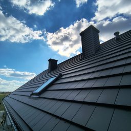 Usługi dekarskie Nowakowski - Czarny dach z dachówką modułową, lukarną i kominami na tle błękitnego nieba z chmurami. Widoczne rynny i fragment rusztowania.