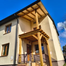 Usługi dekarskie Nowakowski - Dwukondygnacyjny dom z drewnianą konstrukcją zadaszenia nad wejściem, widok z dołu na tle błękitnego nieba. Widoczne rynny i okna.