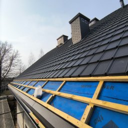 Usługi dekarskie Nowakowski - Czarny dach z kominami w trakcie montażu, widoczne drewniane elementy konstrukcji i niebieska membrana. Poziomica na krawędzi dachu. Ujęcie z dołu.