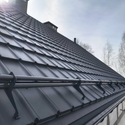 Usługi dekarskie Nowakowski - Grafitowy dach z metalowej dachówki z systemem przeciwśniegowym, widok z dołu, komin z cegły, drzewa w tle, błękitne niebo. Nowoczesne pokrycie dachowe.