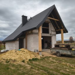 Usługi dekarskie Nowakowski - Dom w trakcie budowy z czarnym dachem i kominem, widoczna więźba dachowa, przyczepka z materiałami budowlanymi i sterta piasku. Pochmurne niebo w tle.