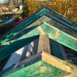 Usługi dekarskie Nowakowski - Wysoki kąt na fragment drewnianej więźby dachowej z zielonymi elementami, widoczne połączenia śrubowe i fragment krajobrazu z drzewami w tle.