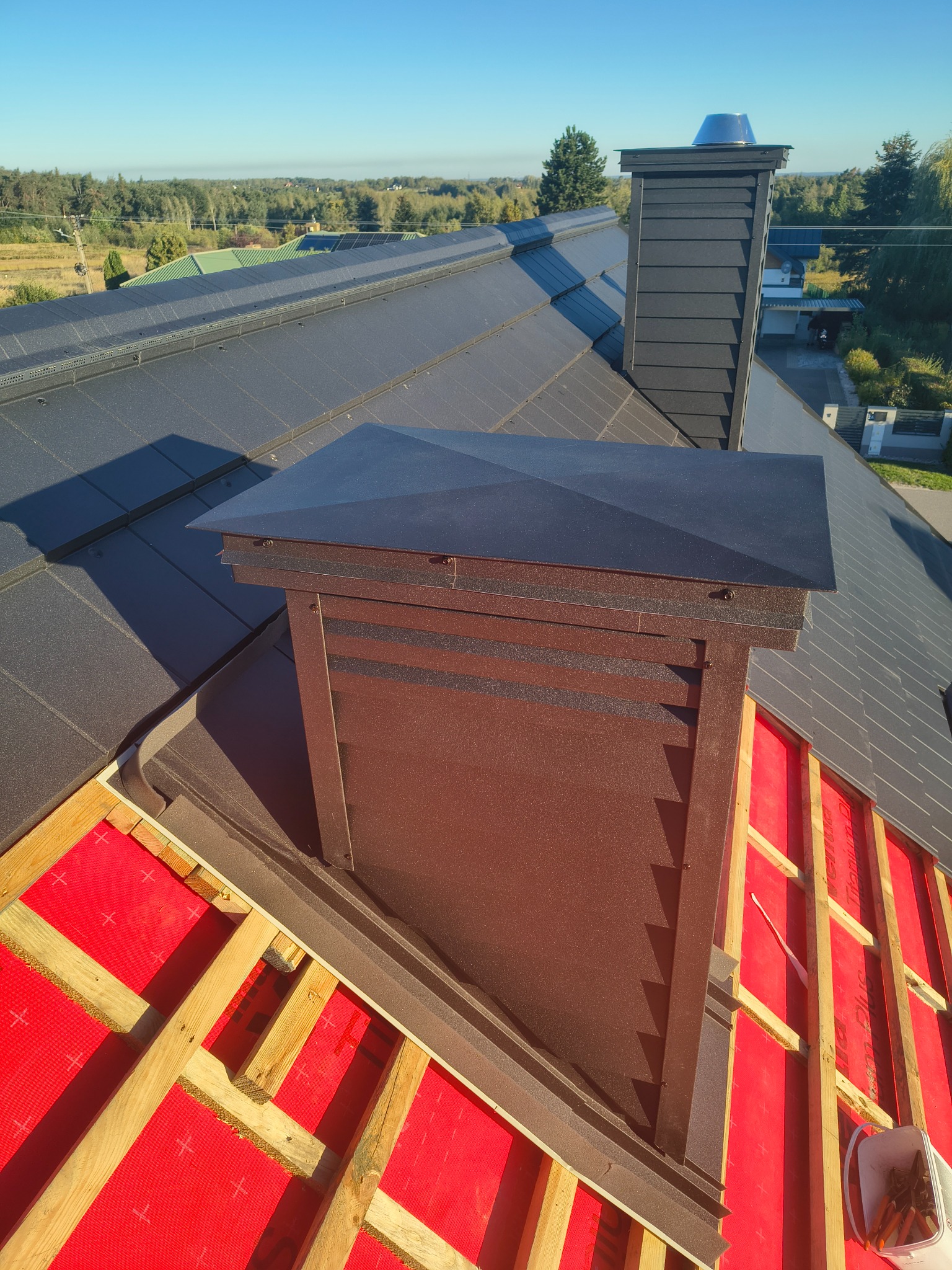 Nowy, ciemny dach z kominem w trakcie budowy. Widoczna czerwona membrana i drewniana konstrukcja. Ujęcie z góry, w tle zieleń drzew i błękitne niebo.