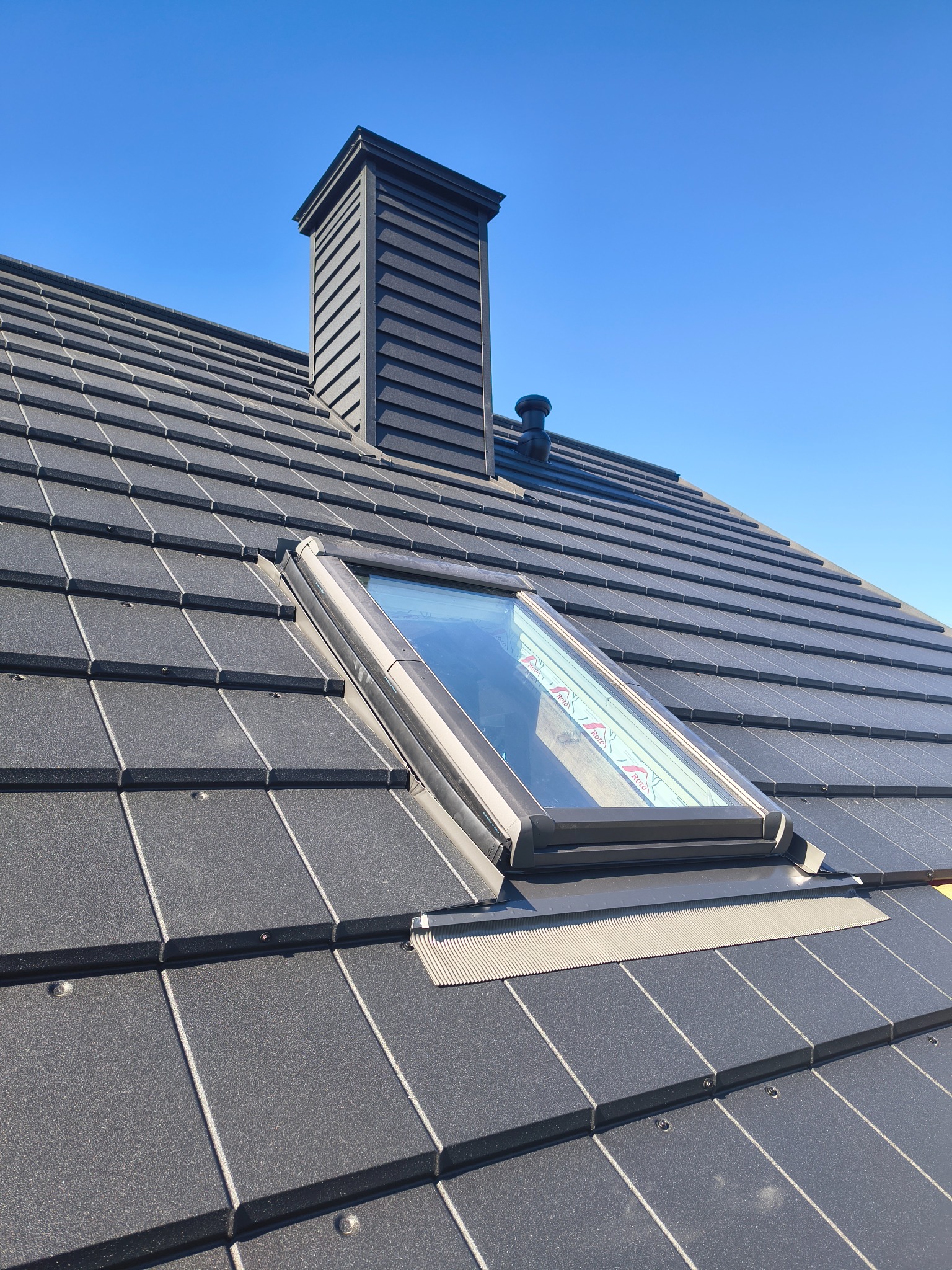 Grafitowy dach z dachówką ceramiczną, kominem i oknem połaciowym w słoneczny dzień. Widok z dołu, podkreślający regularność wzoru dachówki i detale wykończenia.