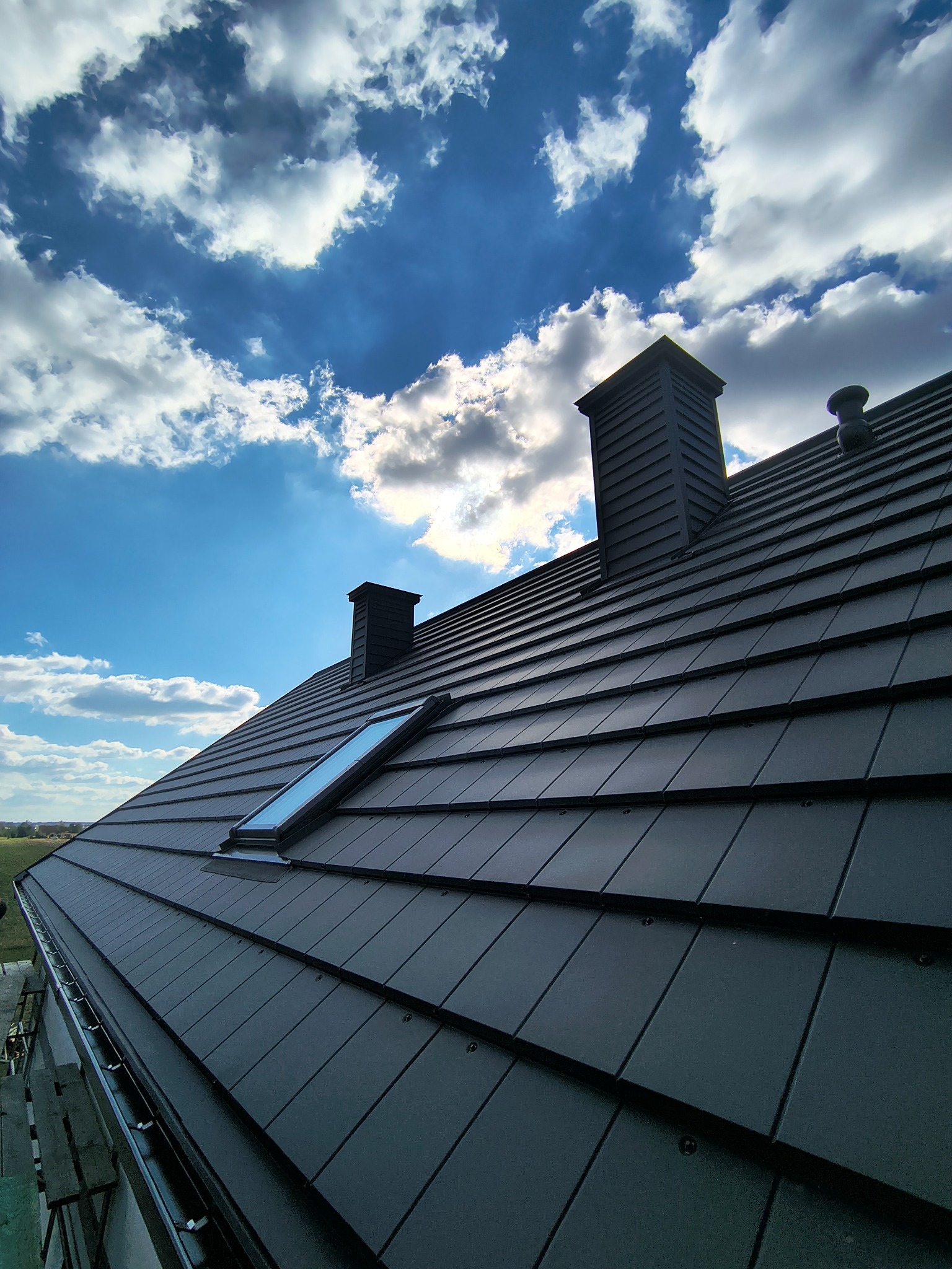 Czarny dach z dachówką modułową, lukarną i kominami na tle błękitnego nieba z chmurami. Widoczne rynny i fragment rusztowania.
