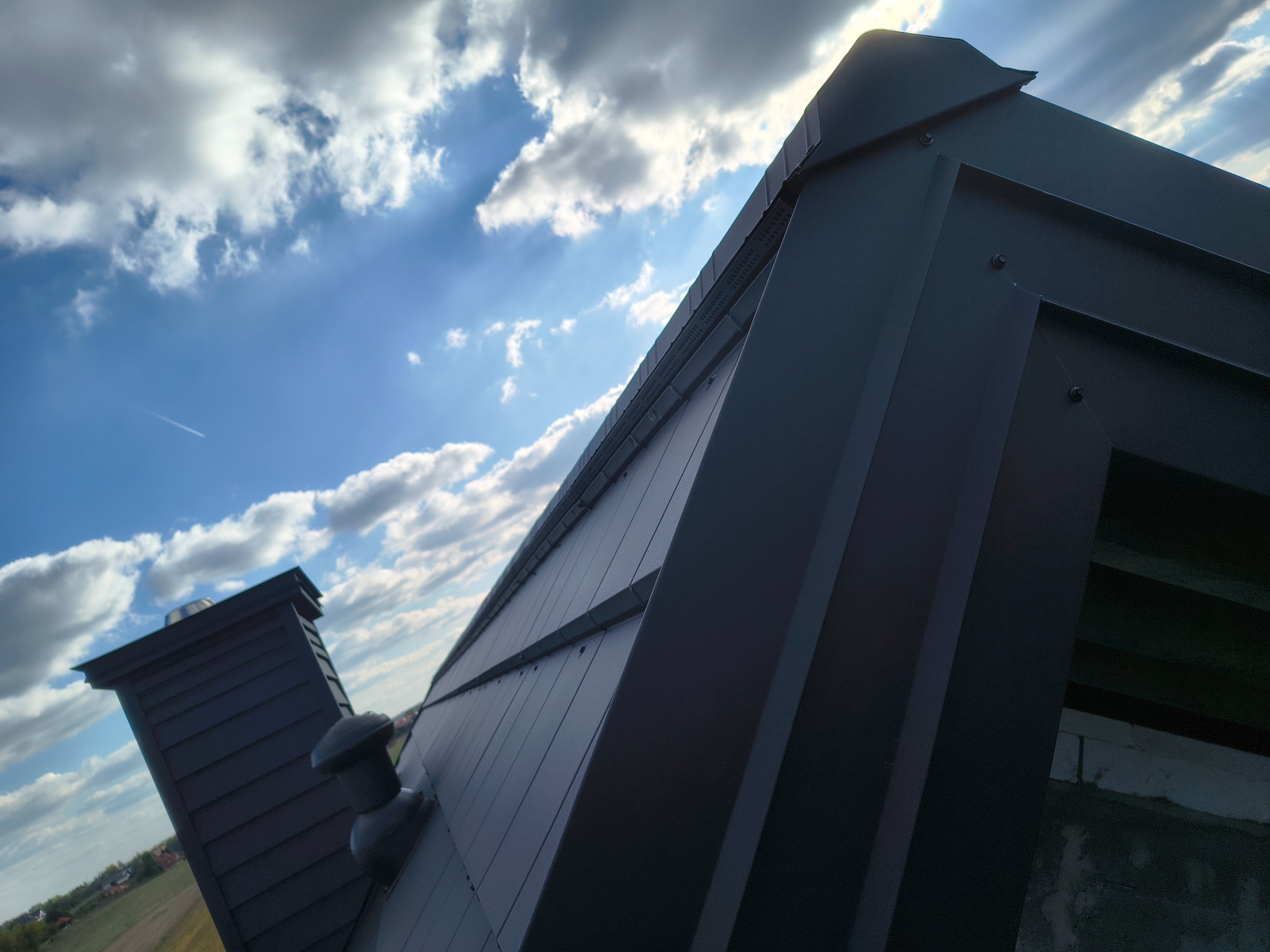 Czarny, metalowy dach z kominem pod kątem, na tle błękitnego nieba z chmurami. Widoczny fragment elewacji i pola. Nietypowy kadr.