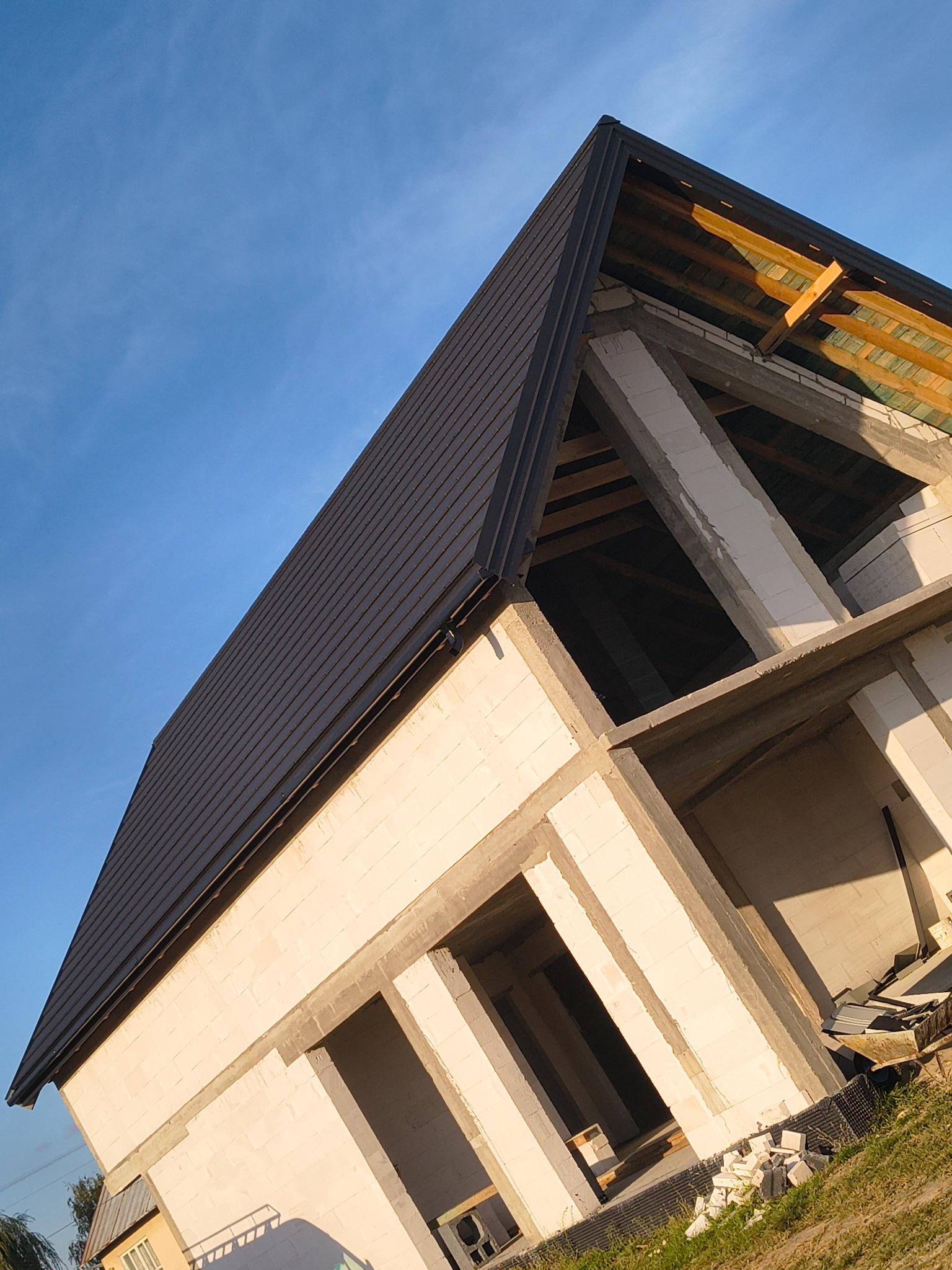 Niski kąt ujęcia domu w trakcie budowy z ciemnym dachem z blachy trapezowej i widoczną więźbą dachową. Białe ściany z betonu komórkowego w surowym stanie.