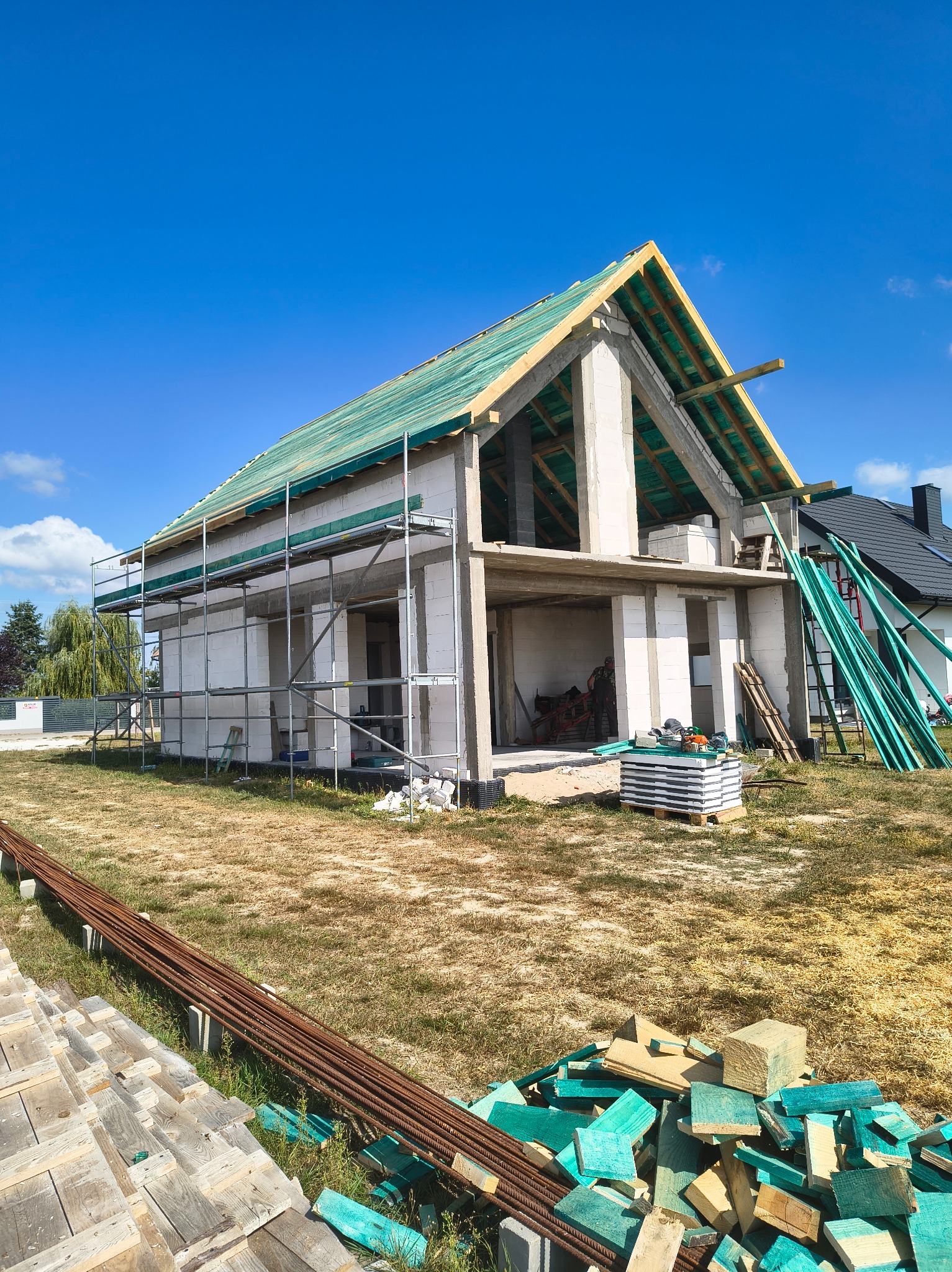 Budowa domu jednorodzinnego w stanie surowym otwartym, z widoczną więźbą dachową pokrytą folią, rusztowaniami i materiałami budowlanymi na działce.