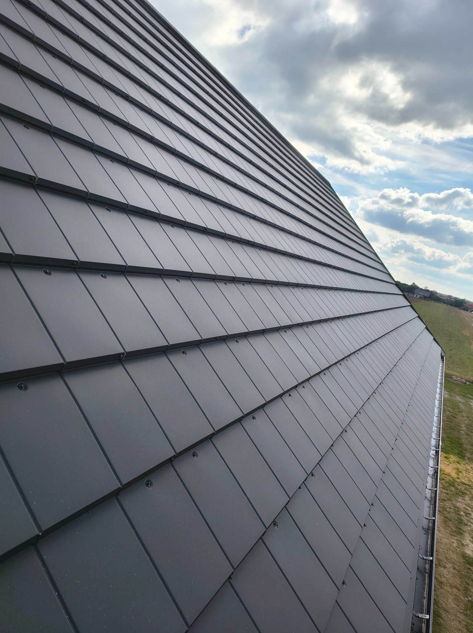 Nowoczesny, grafitowy dach z blachy na rąbek stojący, widok z dołu pod kątem. Na tle błękitne niebo z chmurami i fragment zielonego terenu.