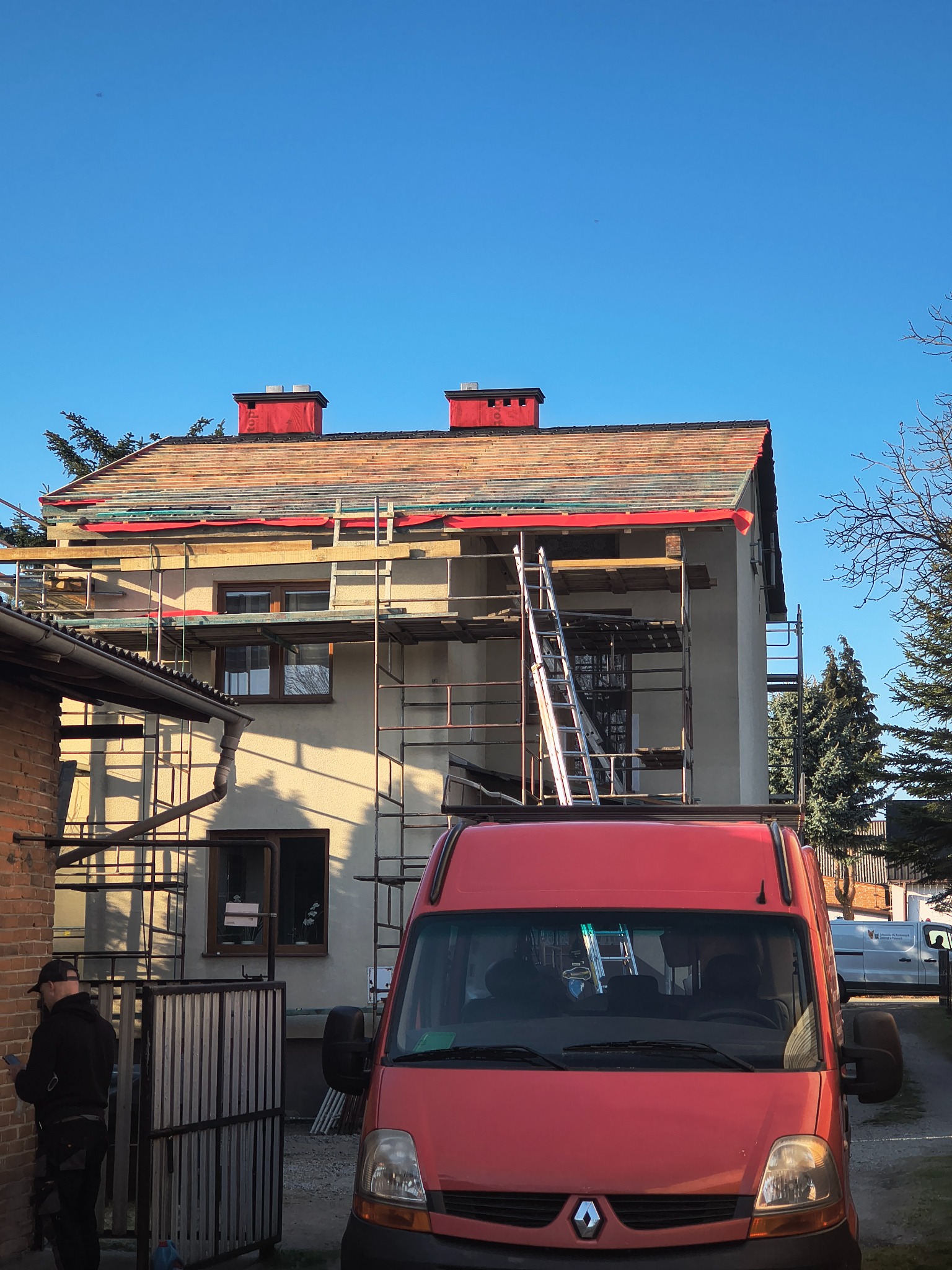 Dom w trakcie wymiany dachu, widoczne rusztowania, częściowo zdjęte stare pokrycie i czerwone kominy. Na pierwszym planie czerwony samochód dostawczy.