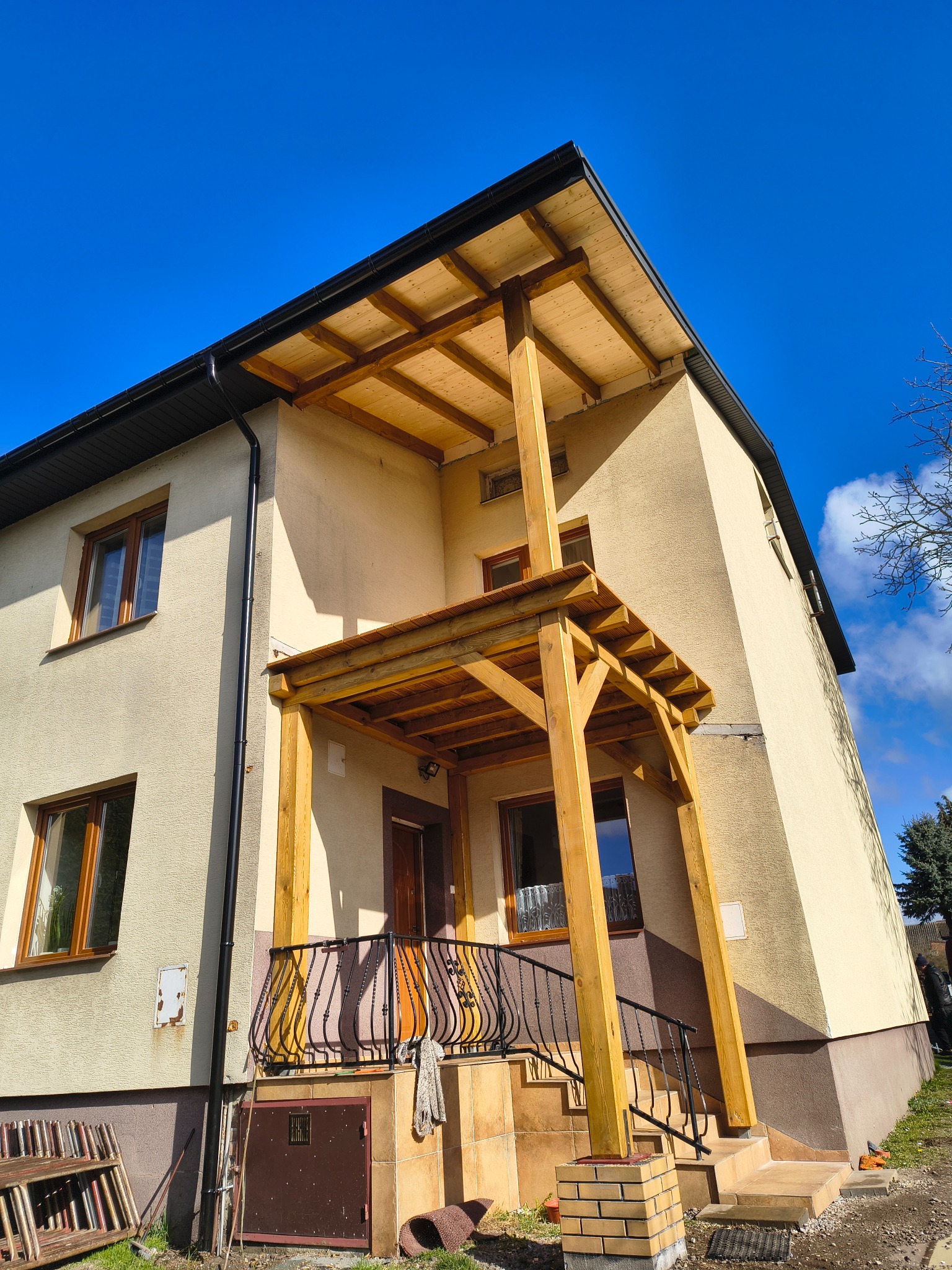 Dwukondygnacyjny dom z drewnianą konstrukcją zadaszenia nad wejściem, widok z dołu na tle błękitnego nieba. Widoczne rynny i okna.