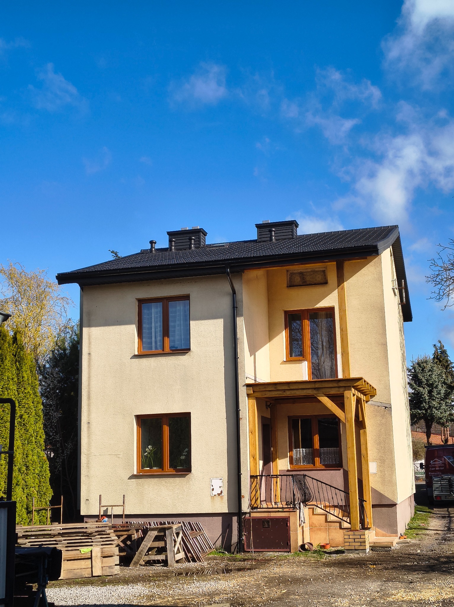 Dwukondygnacyjny dom z nowym, czarnym dachem i drewnianą konstrukcją ganku. Widoczne palety i materiały budowlane. Błękitne niebo z chmurami w tle.