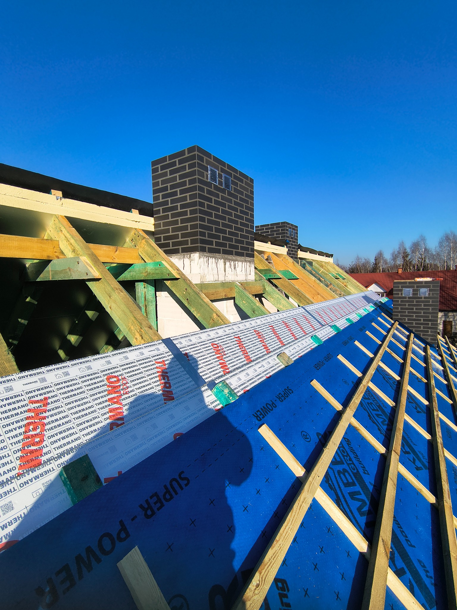 Wysoki kąt na rozłożoną więźbę dachową z membraną i folią izolacyjną, kominy z cegły klinkierowej w tle, błękitne niebo.