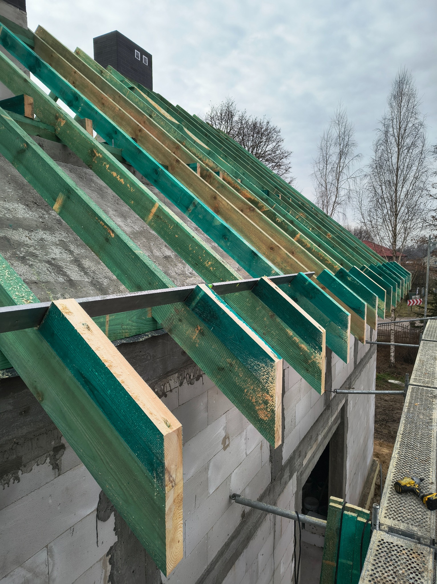 Wznoszenie konstrukcji dachu z impregnowanych belek na budynku z betonu komórkowego. Widoczne rusztowanie i narzędzia. Ujęcie z dołu, pochmurne niebo w tle.