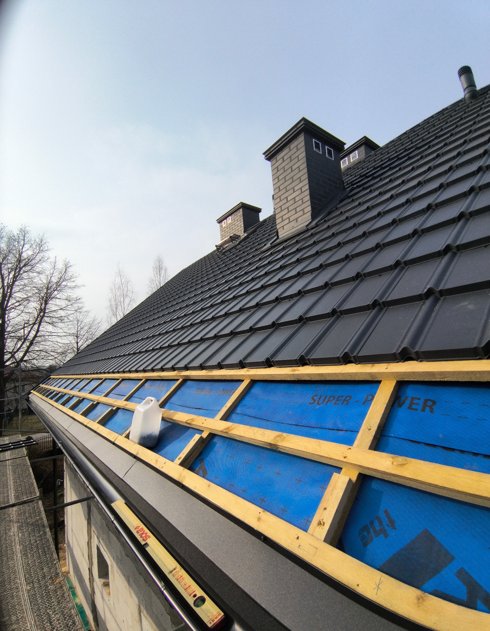 Czarny dach z kominami w trakcie montażu, widoczne drewniane elementy konstrukcji i niebieska membrana. Poziomica na krawędzi dachu. Ujęcie z dołu.