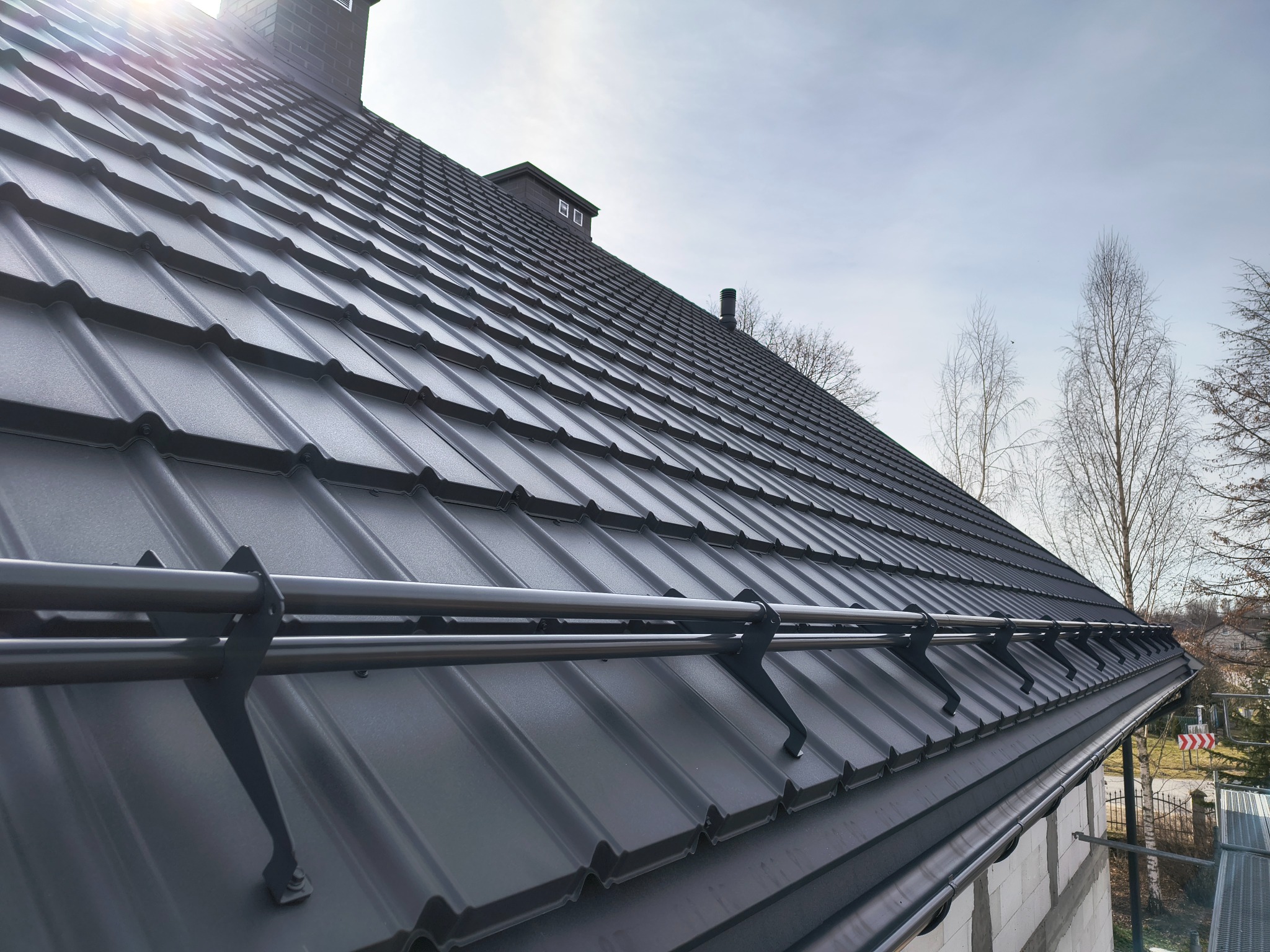 Grafitowy dach z metalowej dachówki z systemem przeciwśniegowym, widok z dołu, komin z cegły, drzewa w tle, błękitne niebo. Nowoczesne pokrycie dachowe.