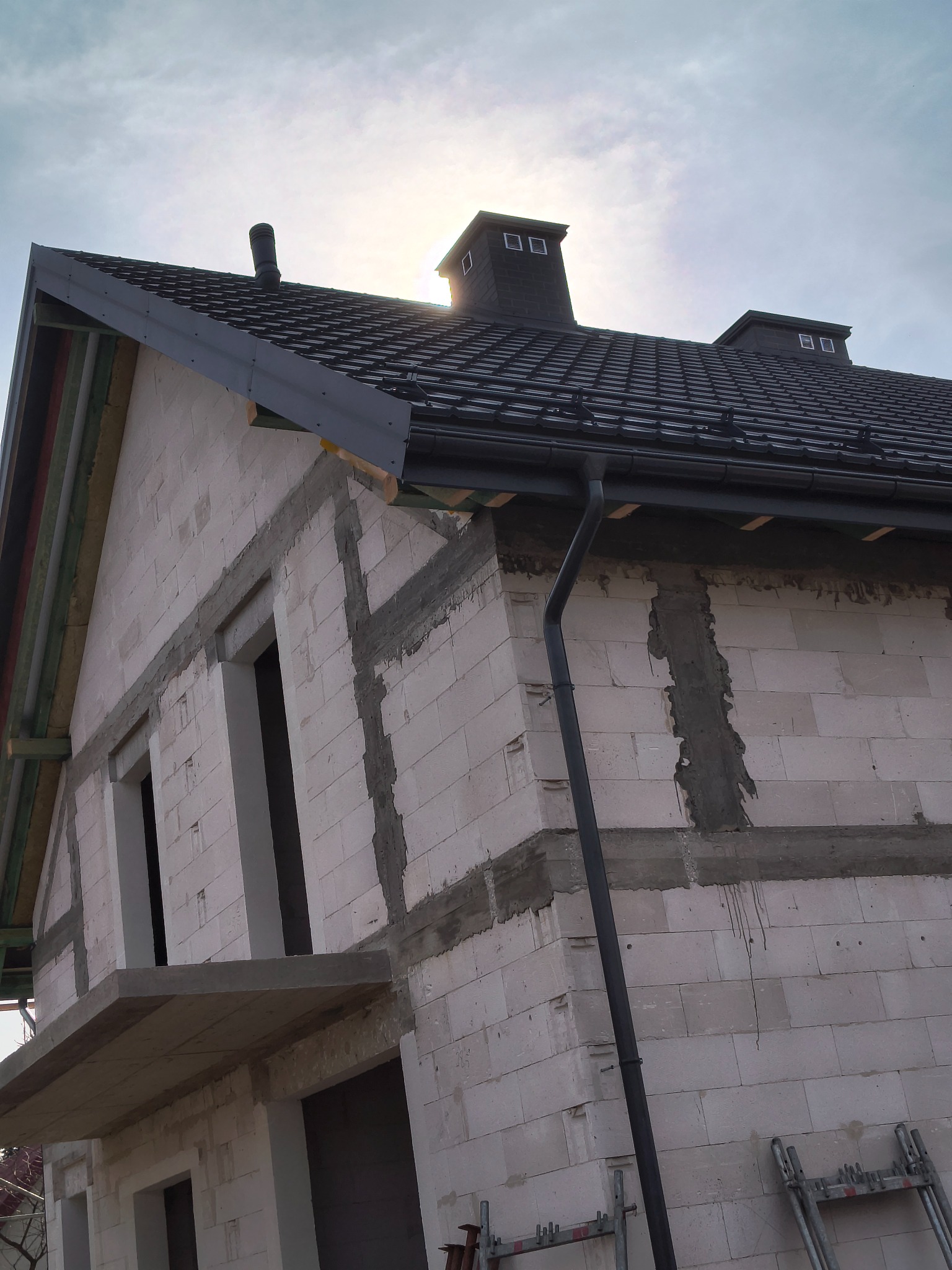 Surowy dom z czarnym dachem i rynnami w trakcie budowy. Widoczne białe bloczki, okna bez szyb i rusztowania. Słońce za dachem.