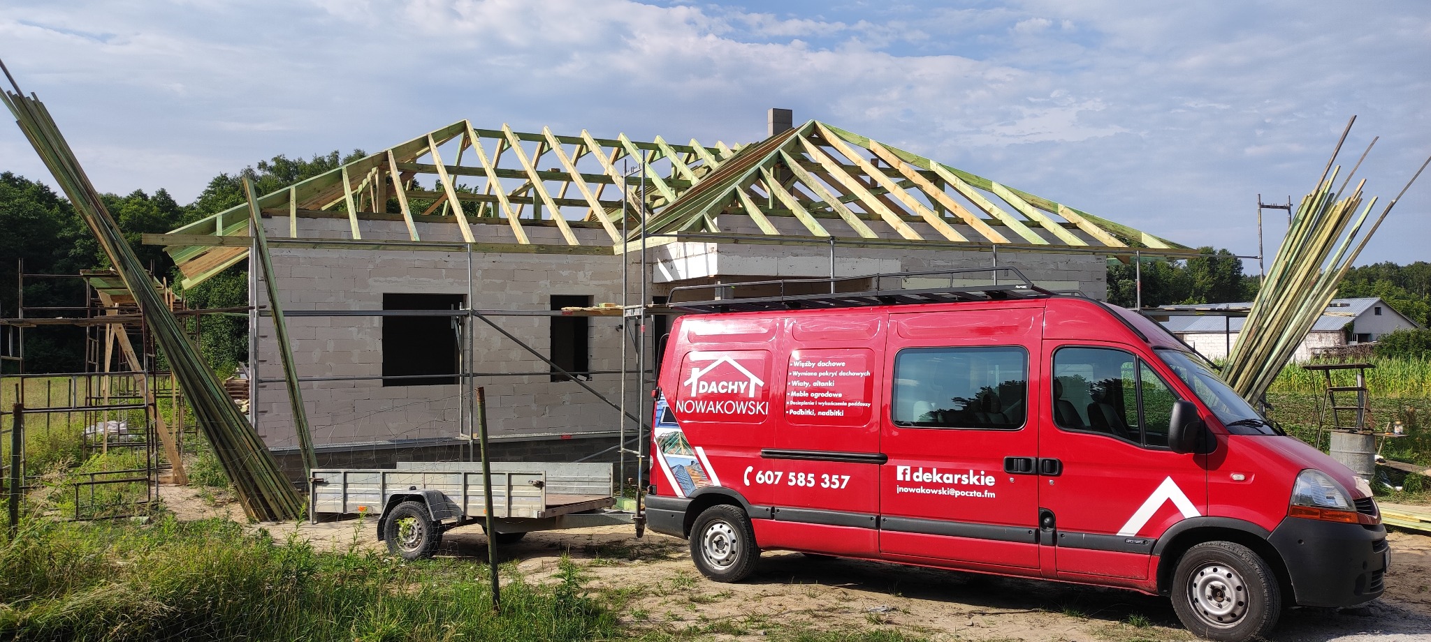 Wznoszona więźba dachowa na budynku w stanie surowym otwartym, z widocznym czerwonym busem firmy dekarskiej i stertą drewna. Prace dekarskie w toku.