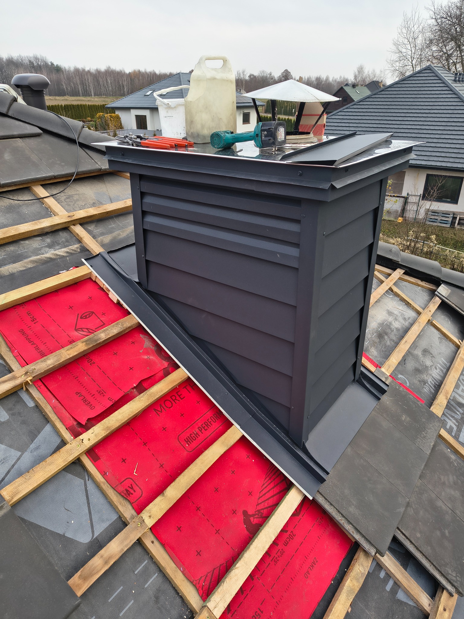 Montaż komina wentylacyjnego na dachu z drewnianą konstrukcją i czerwoną membraną. Widoczne narzędzia i akcesoria dekarskie. Dach pokryty częściowo dachówką.