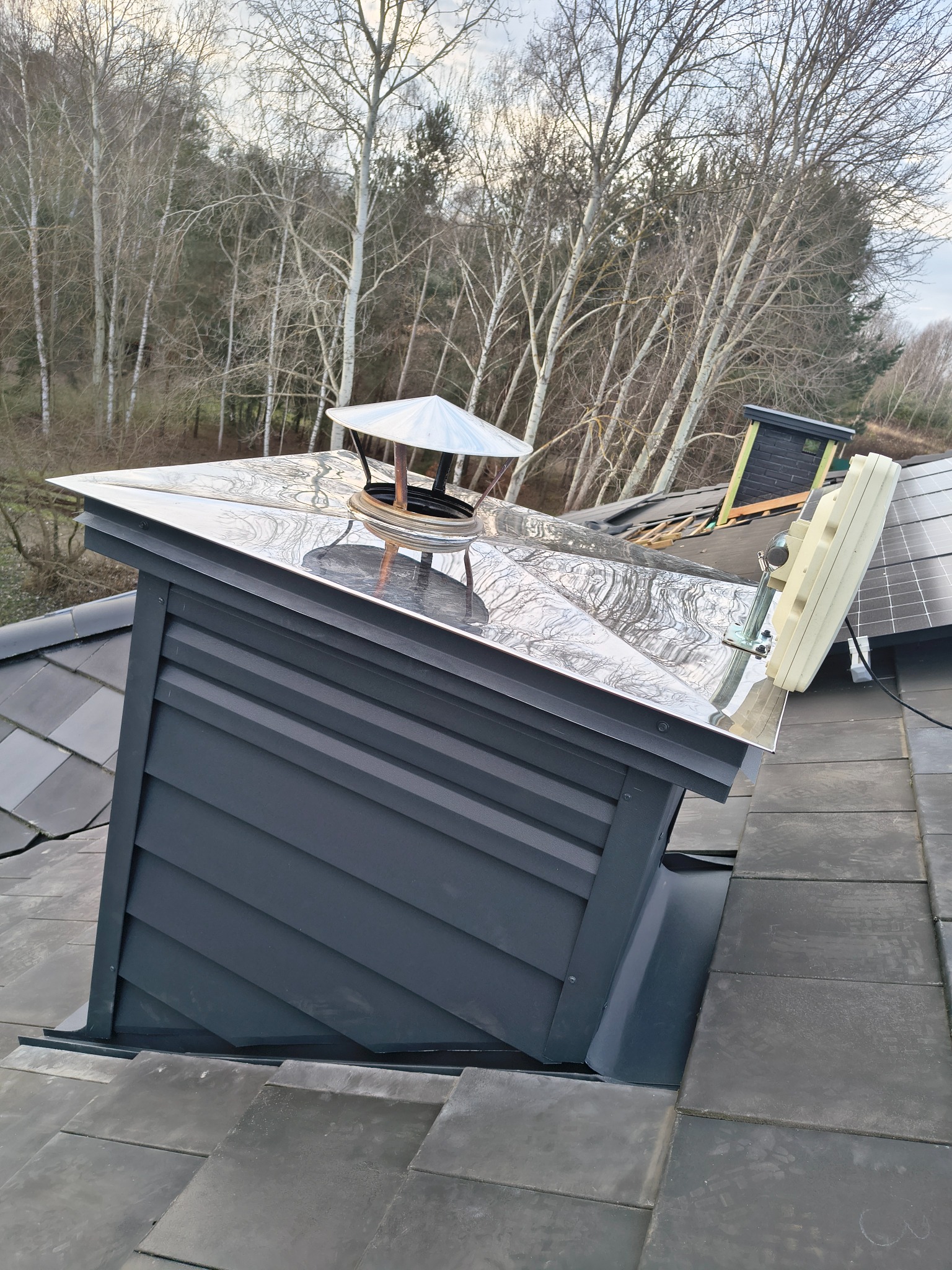 Szczegółowe ujęcie nowego, grafitowego komina z metalowym daszkiem na tle lasu. Widoczne dachówki i elementy instalacji na dachu. Komin z bliska, z wyraźną fakturą.