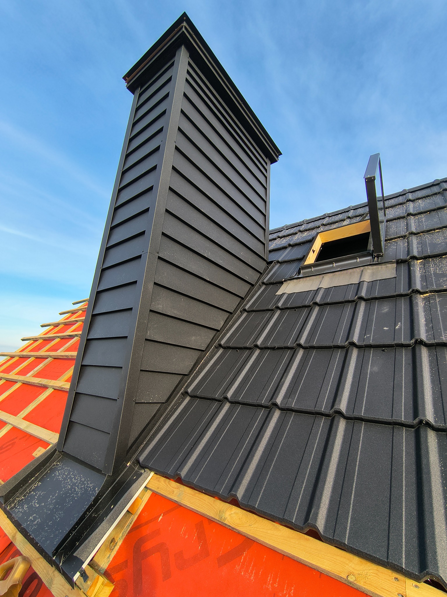 Czarny komin z blachy trapezowej na dachu z dachówki modułowej, widok z dołu na tle błękitnego nieba. Fragment dachu z otwartym oknem dachowym i czerwona membrana dachowa.