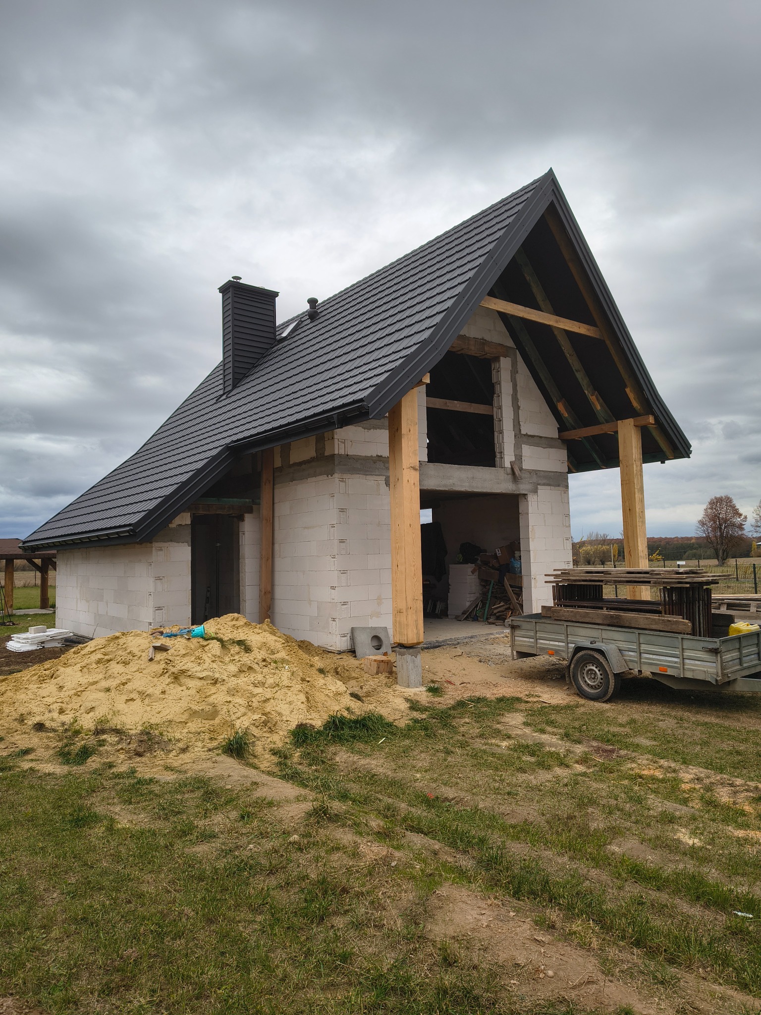 Dom w trakcie budowy z czarnym dachem i kominem, widoczna więźba dachowa, przyczepka z materiałami budowlanymi i sterta piasku. Pochmurne niebo w tle.
