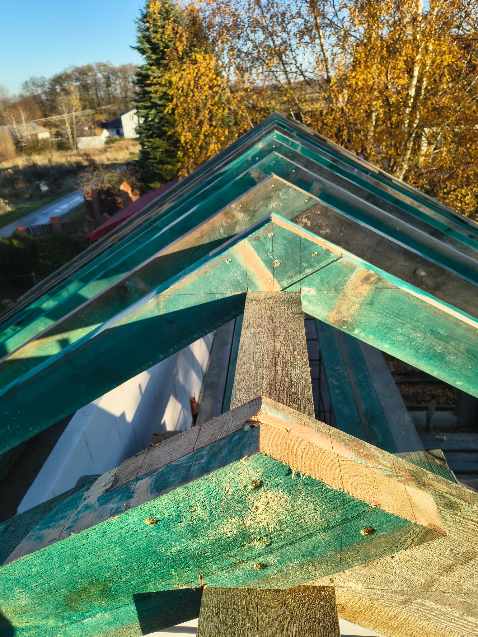 Wysoki kąt na fragment drewnianej więźby dachowej z zielonymi elementami, widoczne połączenia śrubowe i fragment krajobrazu z drzewami w tle.