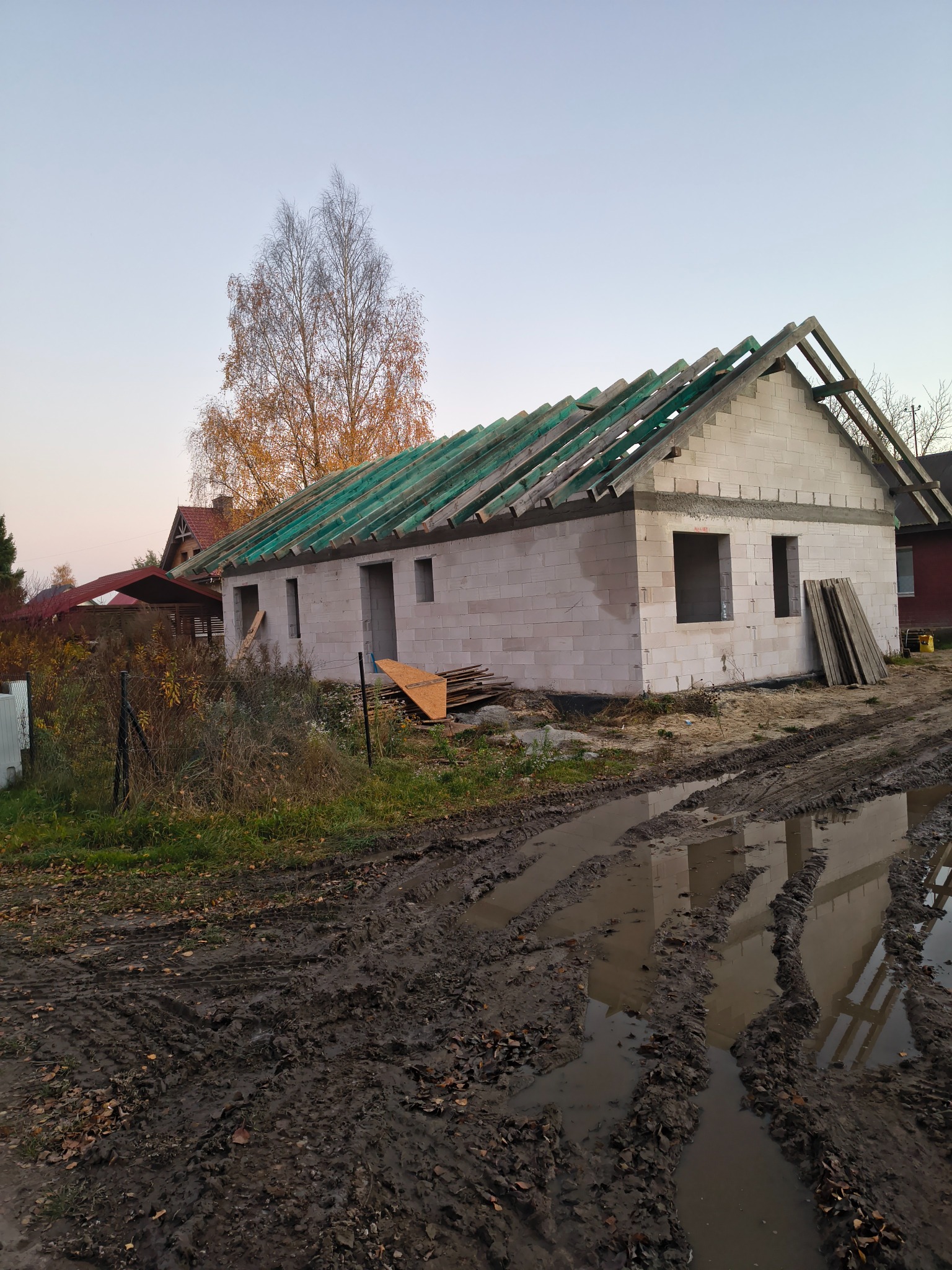 Budowa domu jednorodzinnego w stanie surowym otwartym z widoczną więźbą dachową i elementami konstrukcyjnymi dachu. Błotniste podłoże.