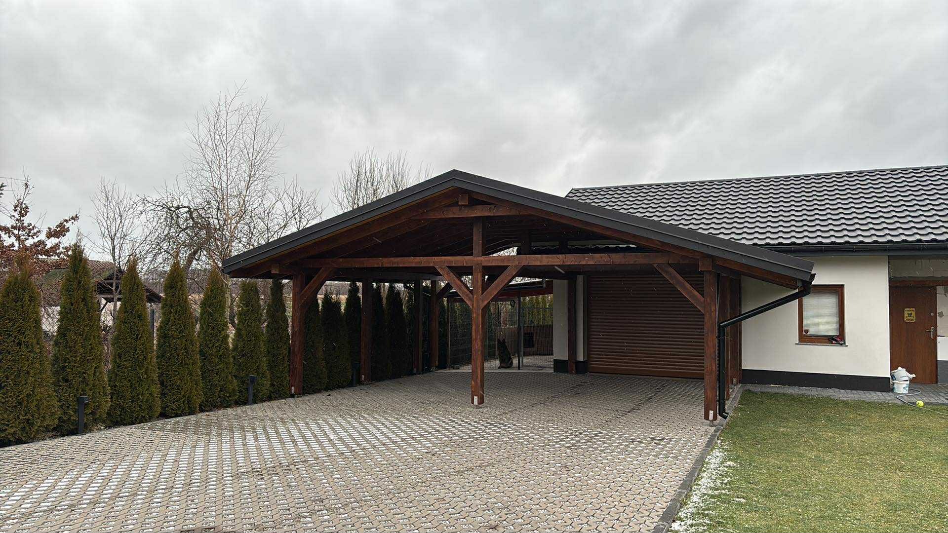 Drewniana wiata garażowa z ciemnym dachem na posesji z kostką brukową i tujami. Widoczny fragment domu z szarą dachówką i rynnami. Pochmurne niebo.