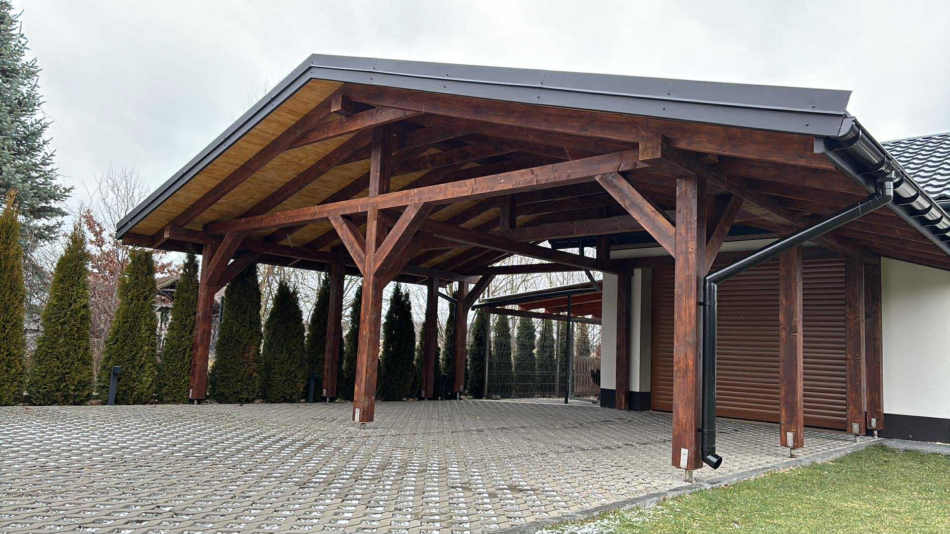 Drewniana wiata garażowa z ciemną, metalową obróbką dachu i rynnami. Widoczne elementy konstrukcyjne, podłoże z ażurowych płyt betonowych i iglaki w tle.