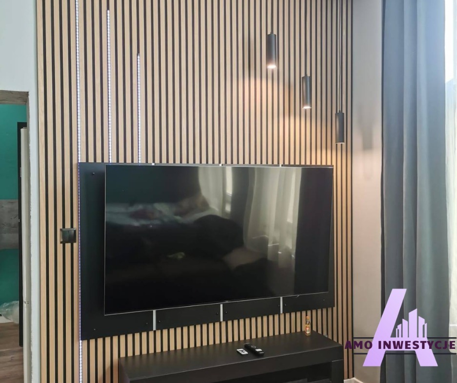 Ściana z lameli z podświetleniem LED, na której wisi telewizor. Nowoczesny design wnętrza z wiszącymi lampami i zasłoną. Widoczny fragment szafki RTV.