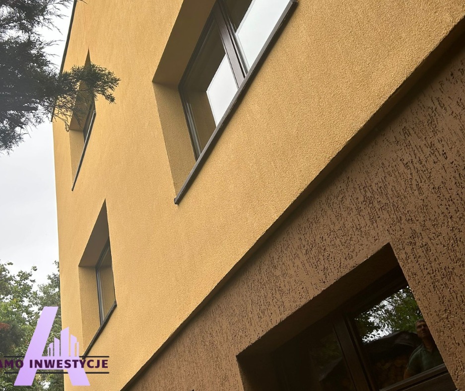 Odnowiona elewacja budynku z oknami, z wyraźnym podziałem na dwa odcienie tynku. Ujęcie z dołu, podkreślające wysokość i strukturę fasady. Widoczny fragment drzewa.