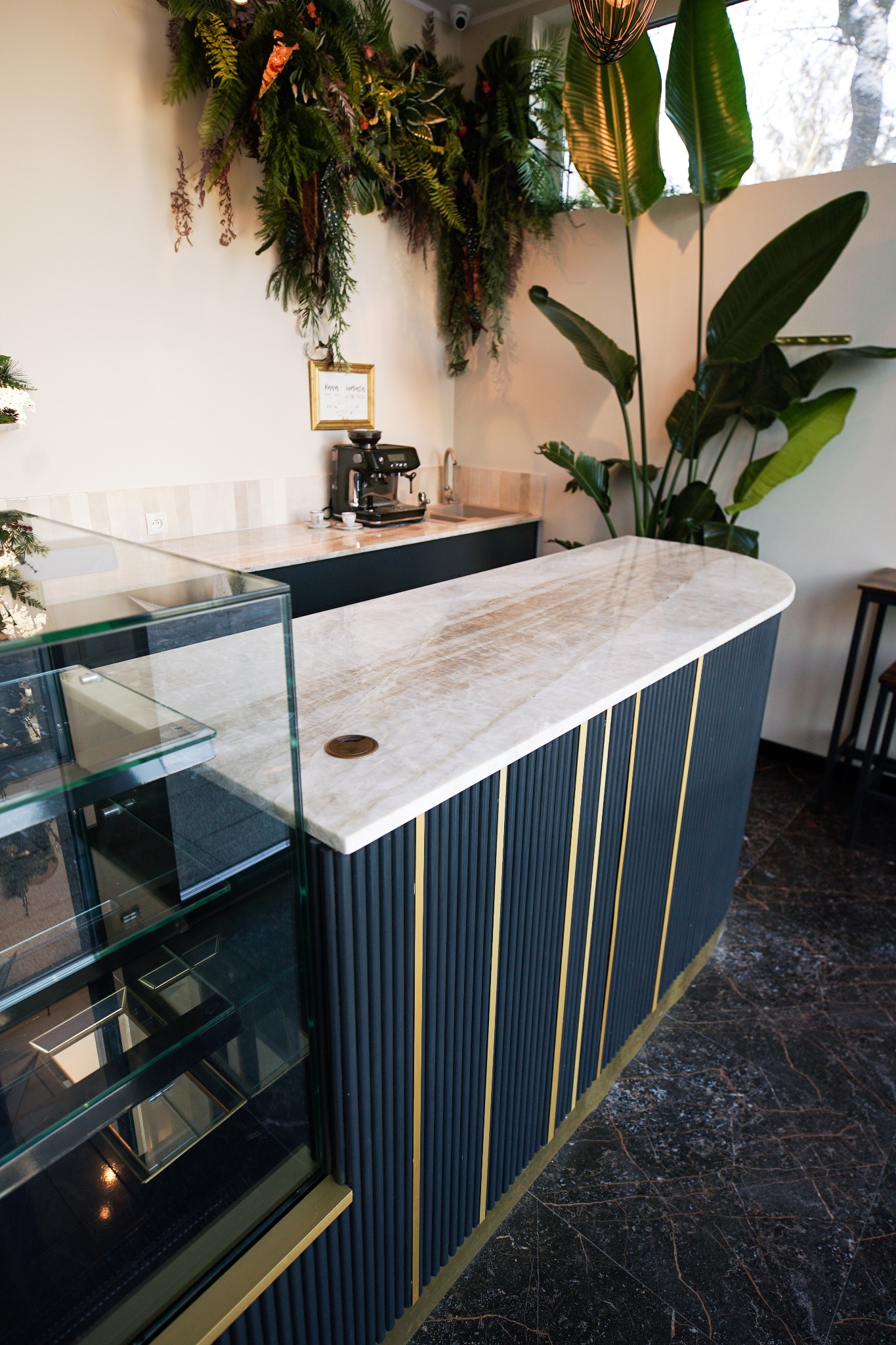 Elegancki bar z marmurowym blatem i ryflowanym frontem w odcieniach granatu i złota, uzupełniony roślinami i ekspozytorem, na tle marmurowej posadzki.