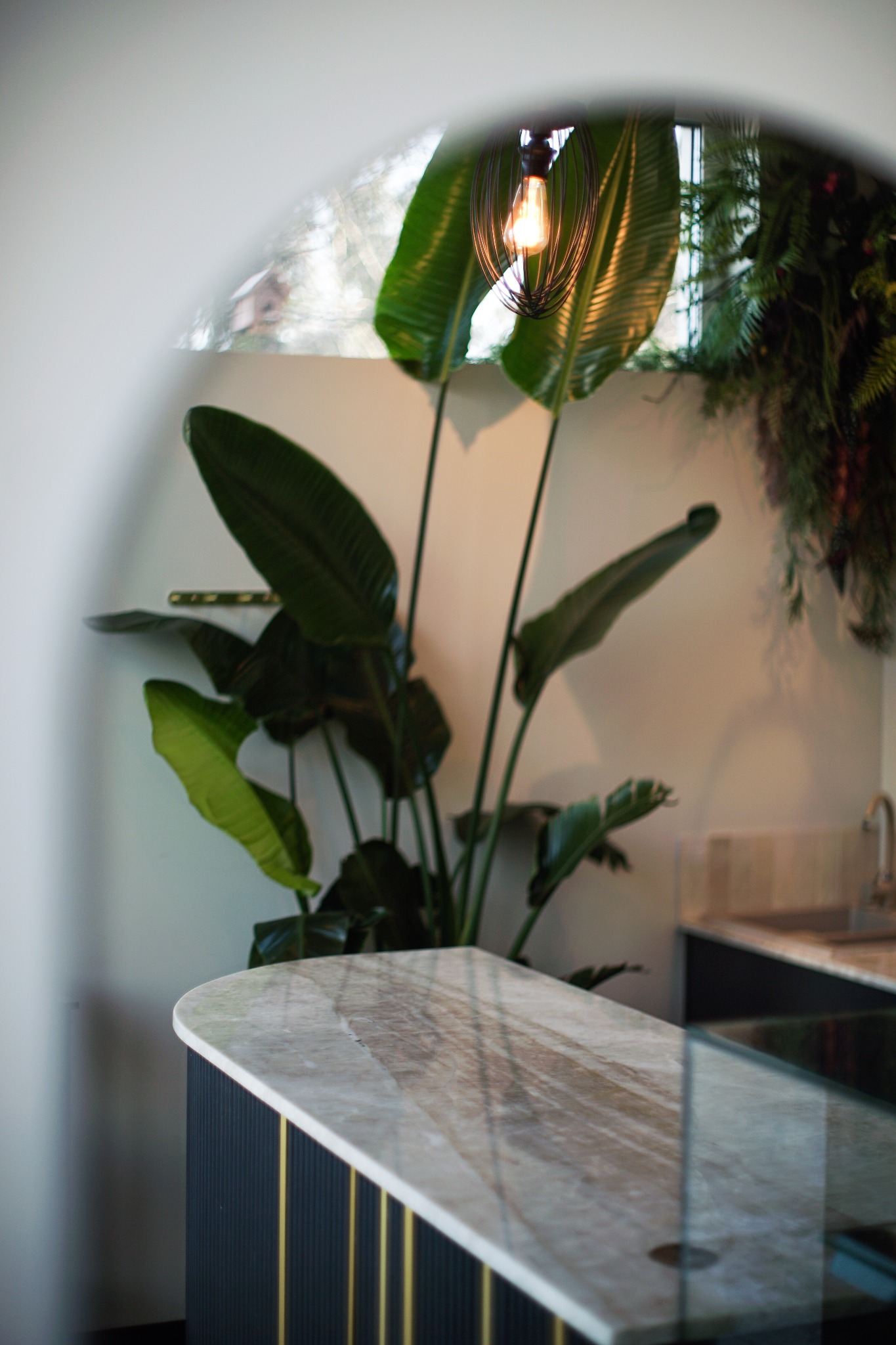 Stylowy blat z marmuru w łazience, oświetlony żarówką w metalowej klatce, z zielonymi roślinami w tle, widziany przez zaokrąglone przejście.