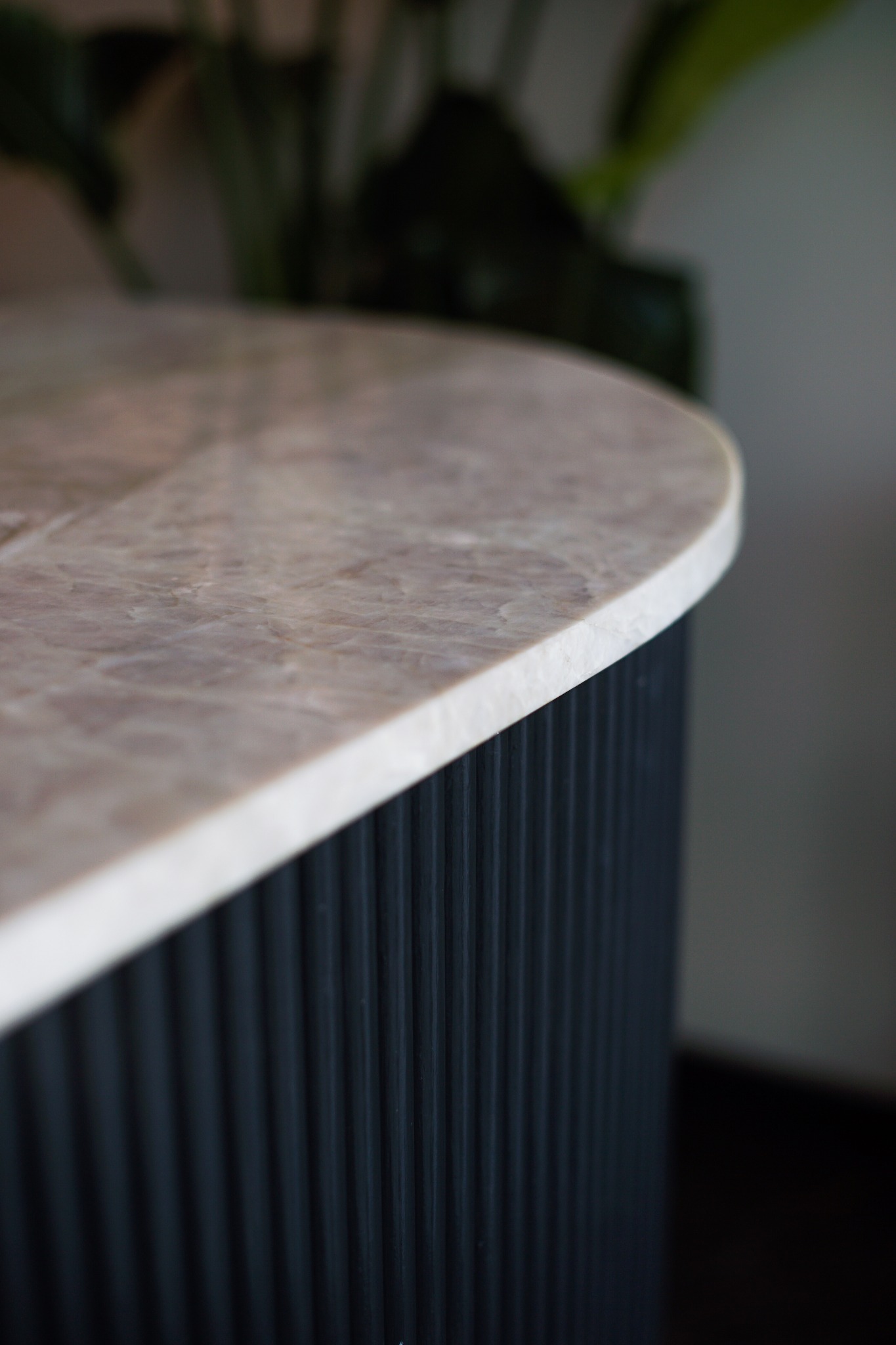 Zaokrąglony blat z beżowego marmuru z białymi żyłkami, wsparty na ciemnogranatowej, ryflowanej podstawie. Fragment rośliny w tle.