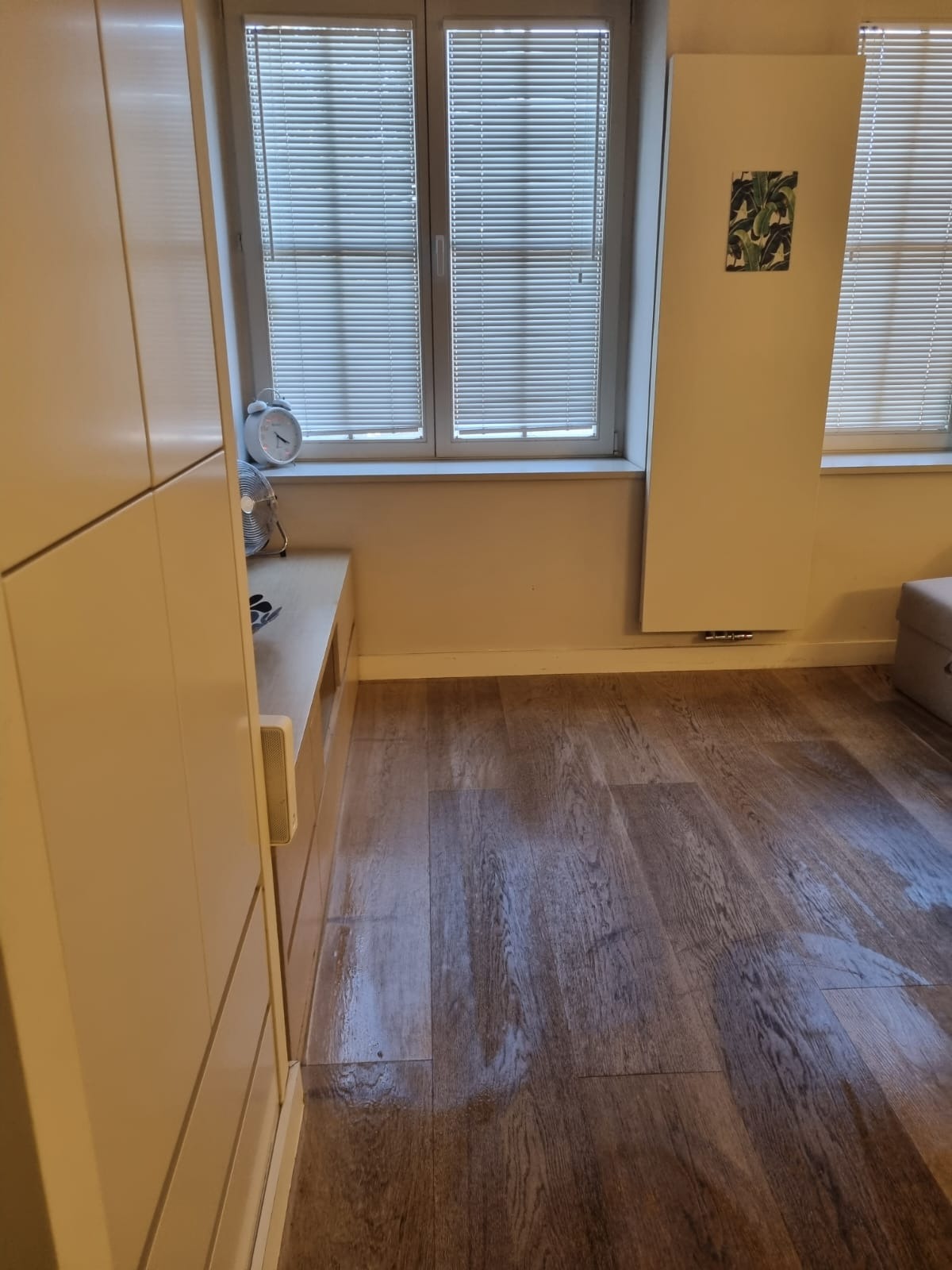 Wnętrze pokoju z drewnianą podłogą, białymi żaluzjami w oknach i jasnymi meblami. Widoczny zegar i wentylator na szafce. Minimalistyczny wystrój.