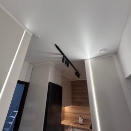 Adrian Przygoda - Nowoczesna kuchnia z białym sufitem podwieszanym, oświetleniem LED wzdłuż ścian i czarną listwą z reflektorami. Widok z dołu, minimalistyczny design.