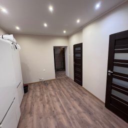 Adrian Przygoda - Jasne wnętrze z panelami podłogowymi w odcieniach brązu, widoczne drzwi wewnętrzne z przeszkleniami i biała szafa. Oświetlenie sufitowe punktowe.