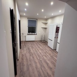 Adrian Przygoda - Jasne wnętrze po remoncie: drewnopodobne panele podłogowe, białe ściany i sufit z punktowym oświetleniem. Widoczne drzwi i fragment kuchni z lodówkami.