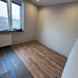 Adrian Przygoda - Częściowo ułożone panele podłogowe w pokoju z oknem. Widoczna podkładka pod panele z niebieską taśmą. Świeżo pomalowane ściany w odcieniach szarości.