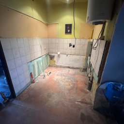 Adrian Przygoda - Demontaż płytek w kuchni przed remontem. Widoczne narzędzia, fragmenty odspojonych płytek i przygotowane podłoże. Ujęcie z perspektywy wejścia do pomieszczenia.