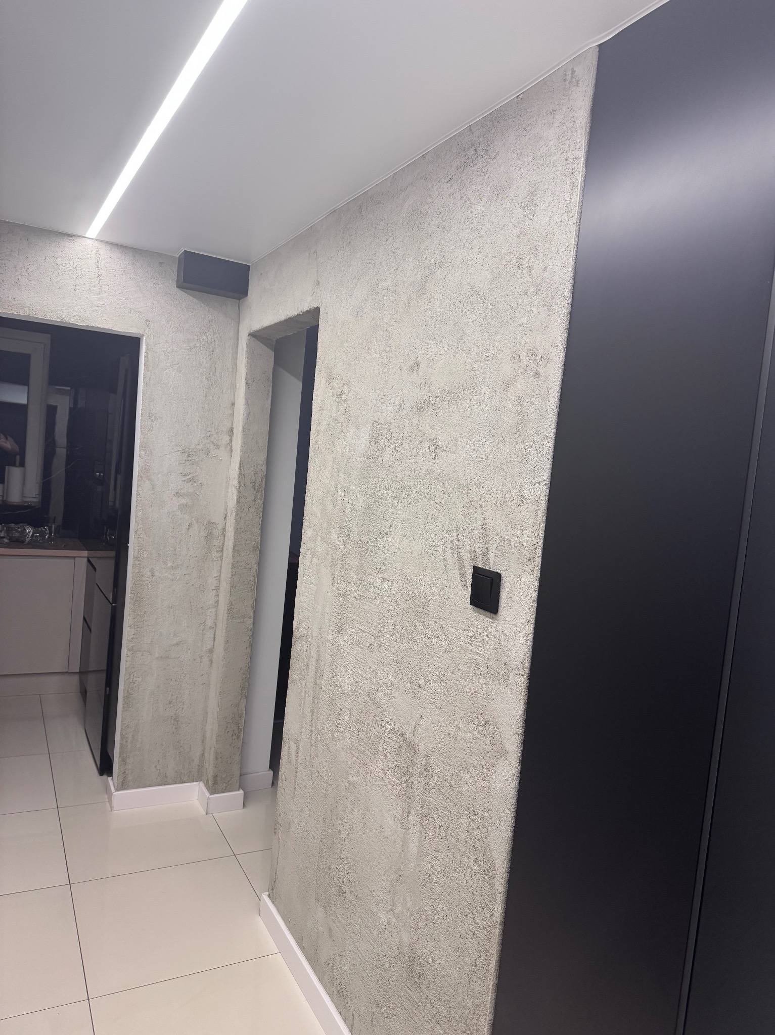Korytarz po odświeżeniu: ściany z szarą, teksturowaną farbą, białe listwy przypodłogowe, jasna podłoga z płytek i czarna szafa w zabudowie. Nowoczesne oświetlenie sufitowe.