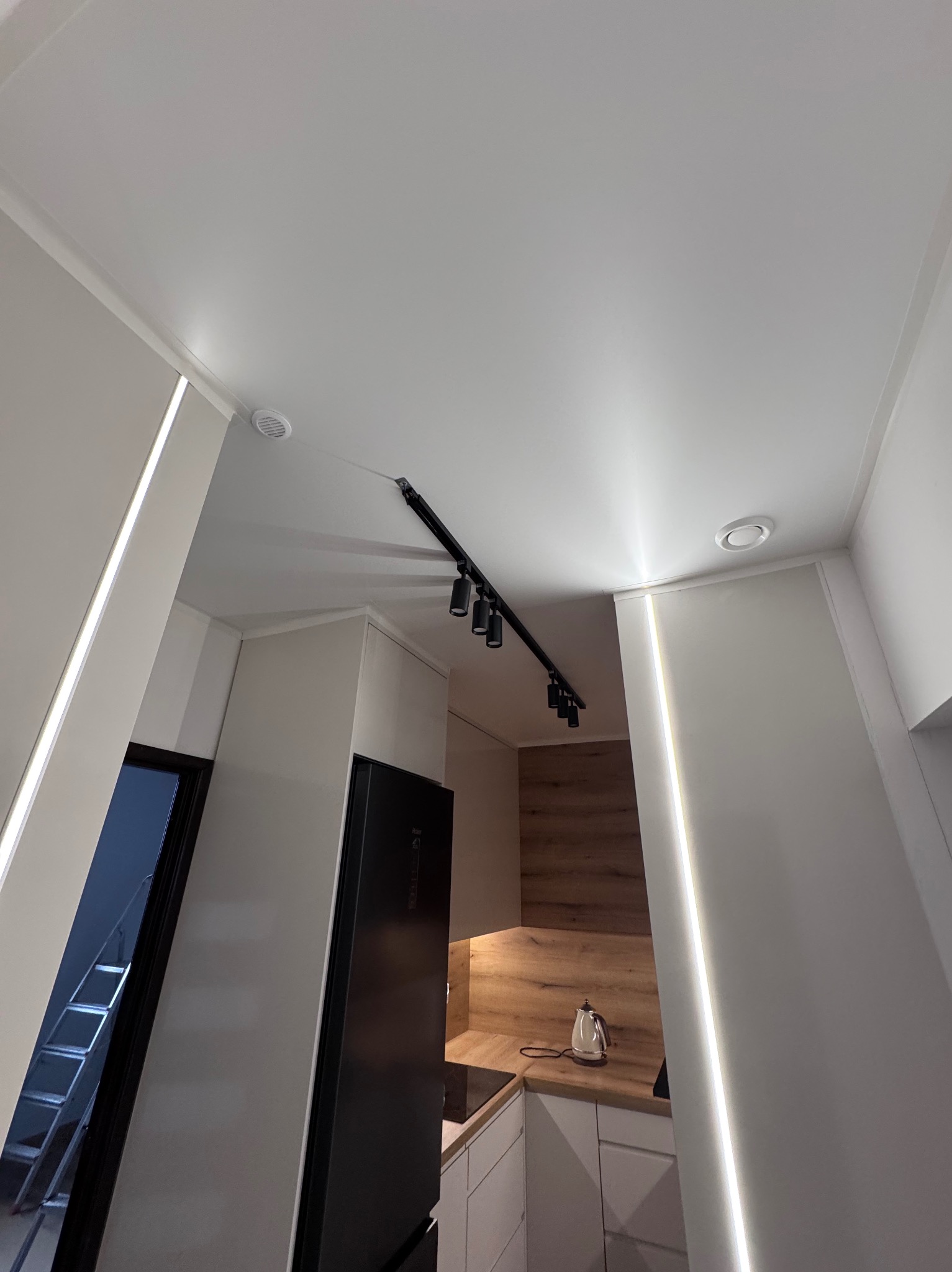 Nowoczesna kuchnia z białym sufitem podwieszanym, oświetleniem LED wzdłuż ścian i czarną listwą z reflektorami. Widok z dołu, minimalistyczny design.