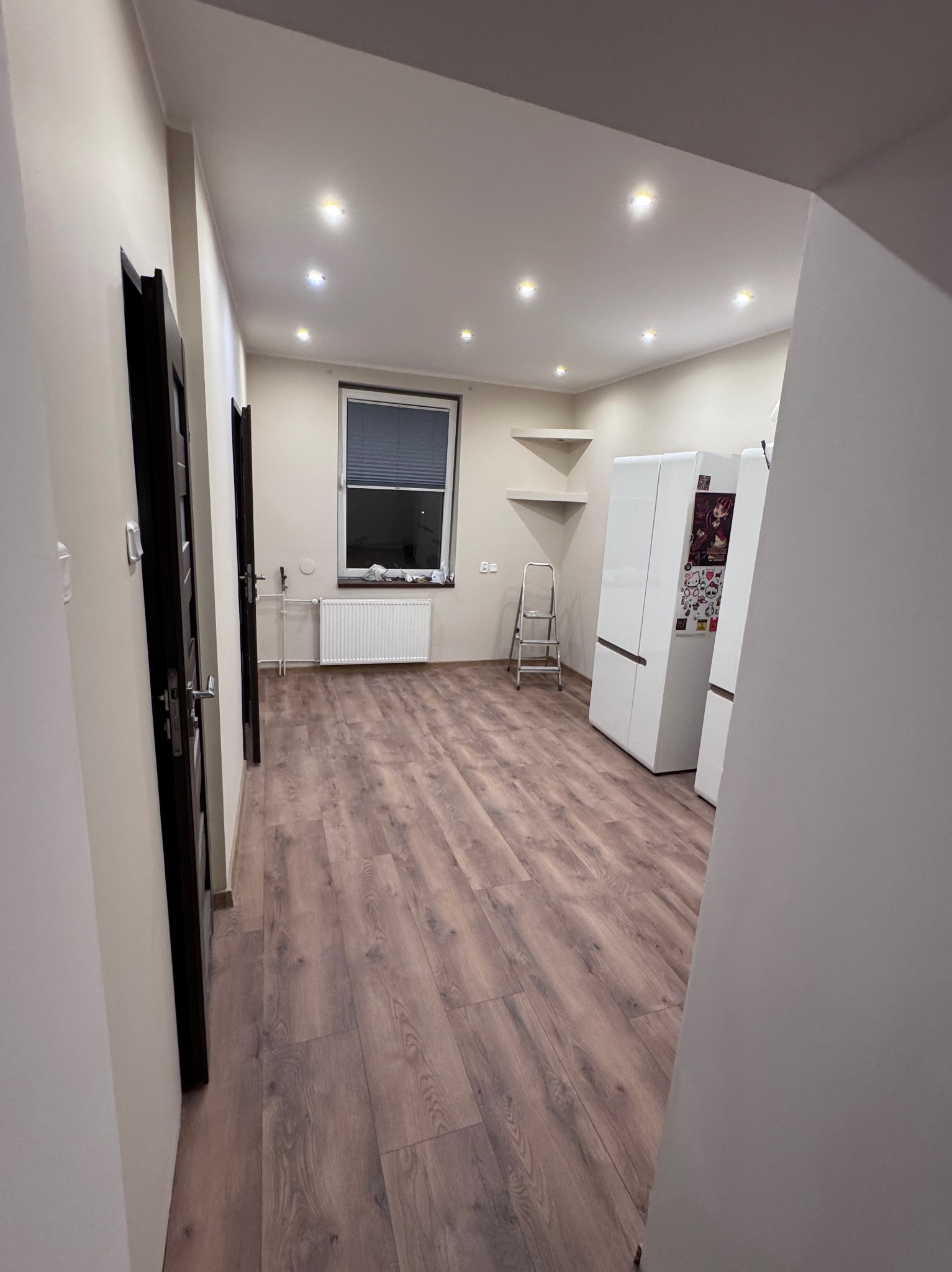 Jasne wnętrze po remoncie: drewnopodobne panele podłogowe, białe ściany i sufit z punktowym oświetleniem. Widoczne drzwi i fragment kuchni z lodówkami.