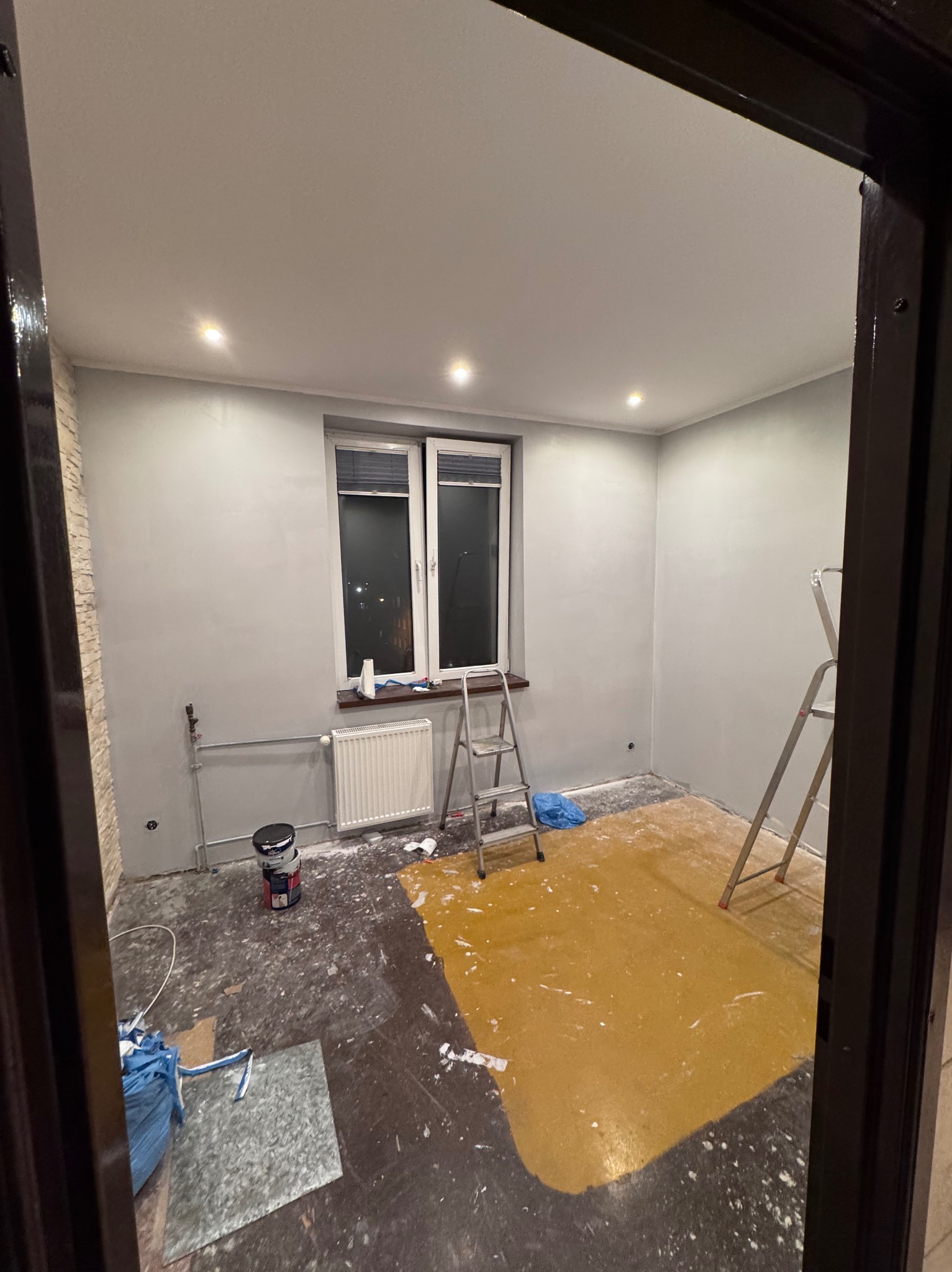 Pokój w trakcie malowania na szaro, widoczne drabiny, puszki z farbą i częściowo pomalowana podłoga na żółto. Widok zza futryny drzwi.