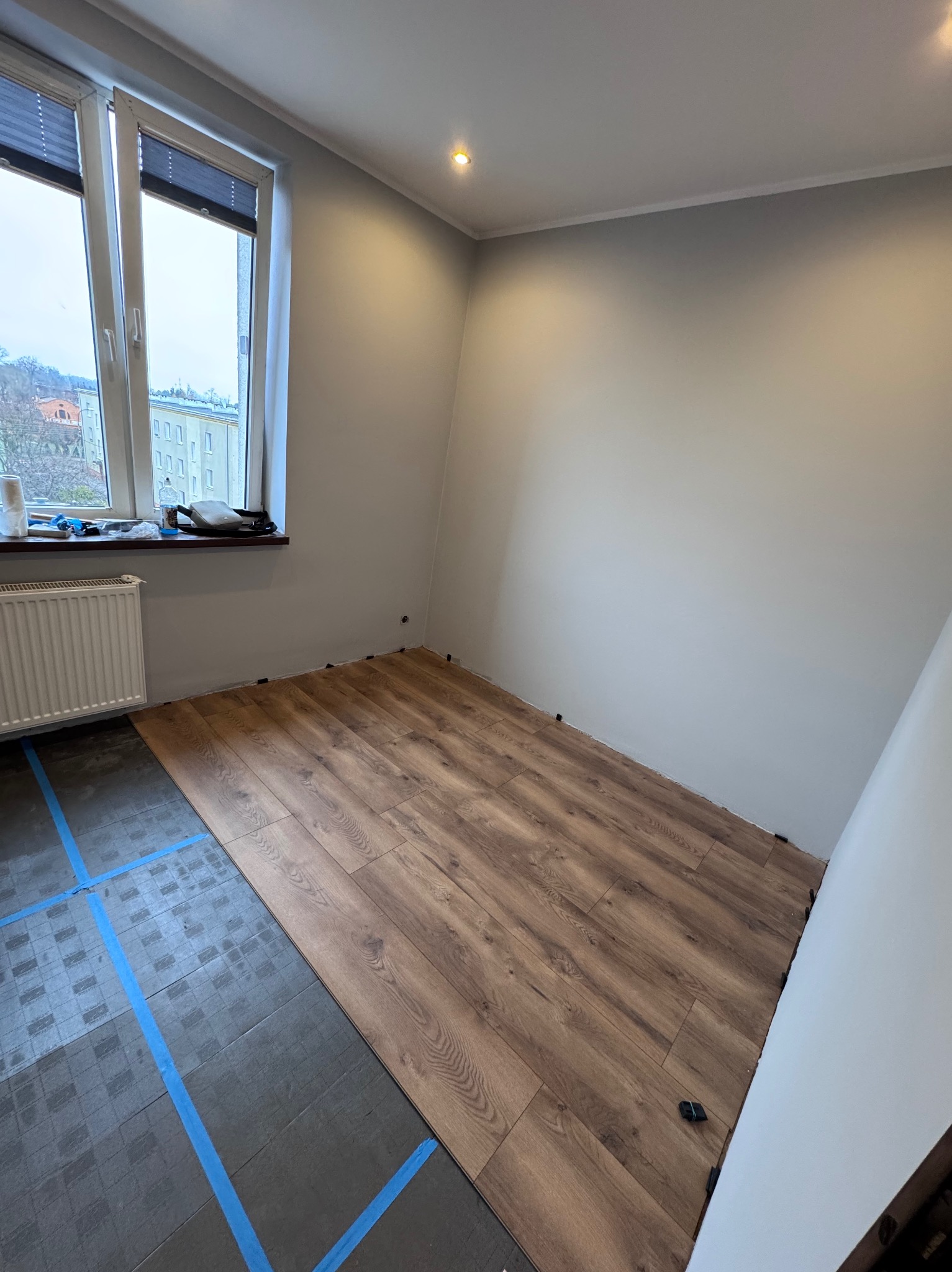Częściowo ułożone panele podłogowe w pokoju z oknem. Widoczna podkładka pod panele z niebieską taśmą. Świeżo pomalowane ściany w odcieniach szarości.