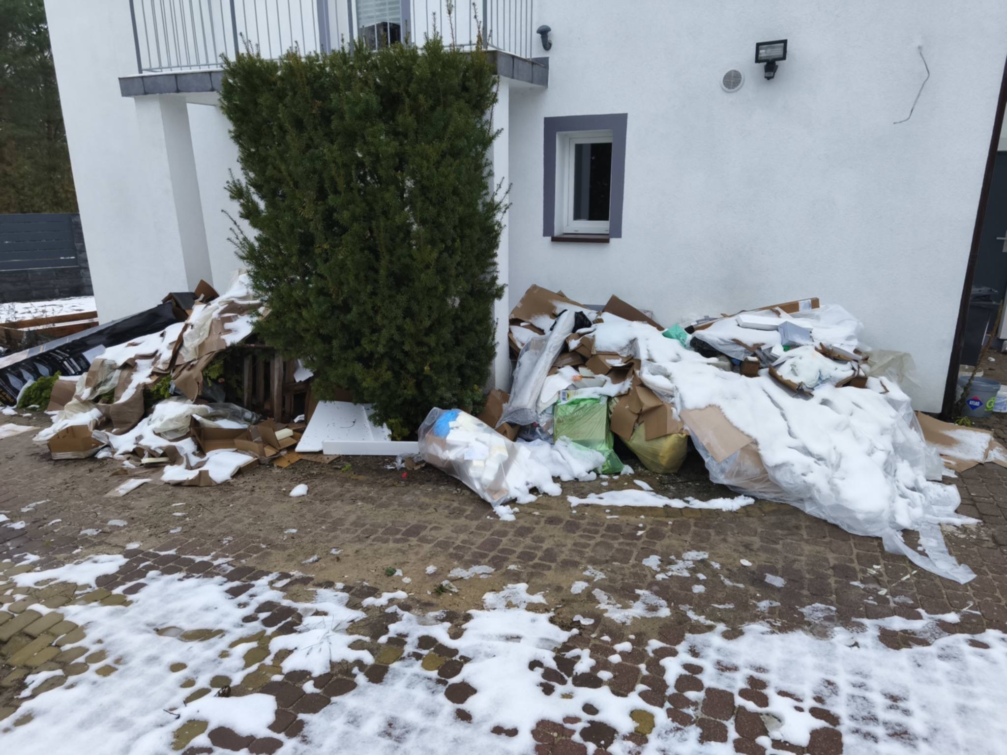 Sterta kartonów, styropianu i worków ze śniegiem przed białym domem z krzewem. Opróżnianie po remoncie w zimowej scenerii.