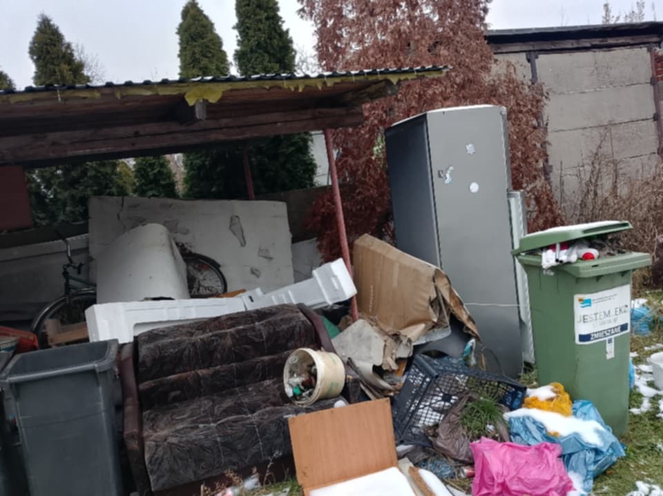Zabudowany śmietnik z meblami, sprzętem AGD i odpadami różnego rodzaju na zewnątrz, z zielonym pojemnikiem na śmieci z boku. Widoczne drzewa w tle.