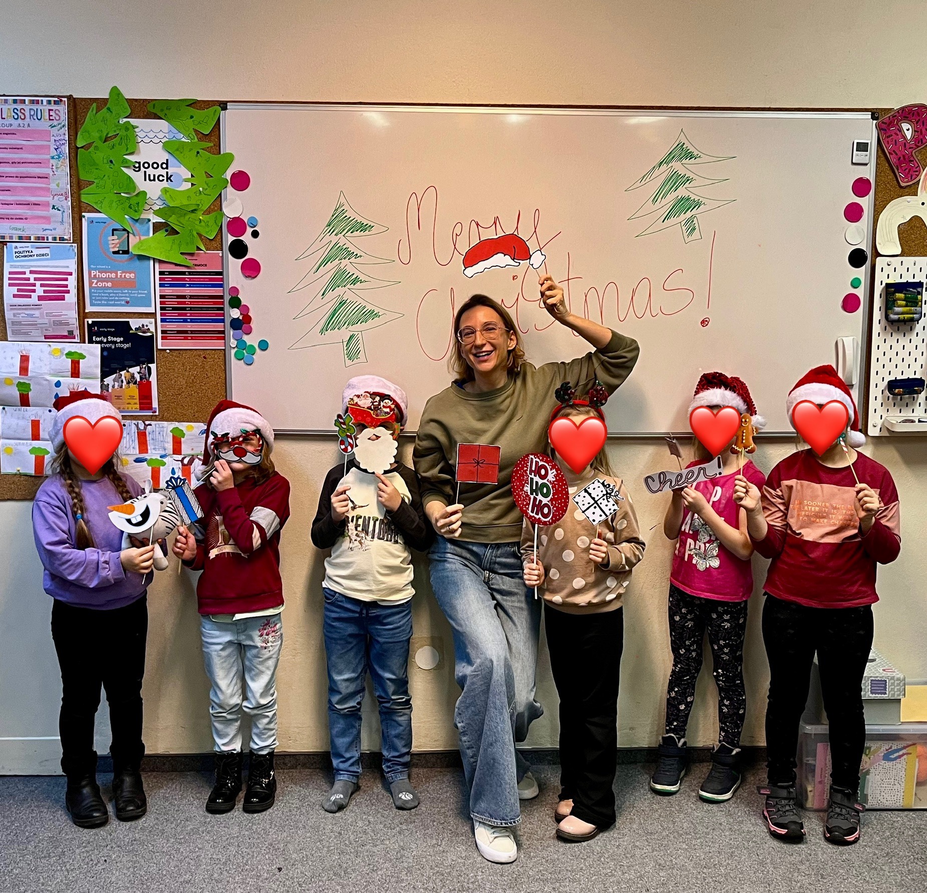 Świąteczna atmosfera w szkole językowej: uśmiechnięta nauczycielka i dzieci z rekwizytami na tle tablicy z napisem 'Merry Christmas!'.