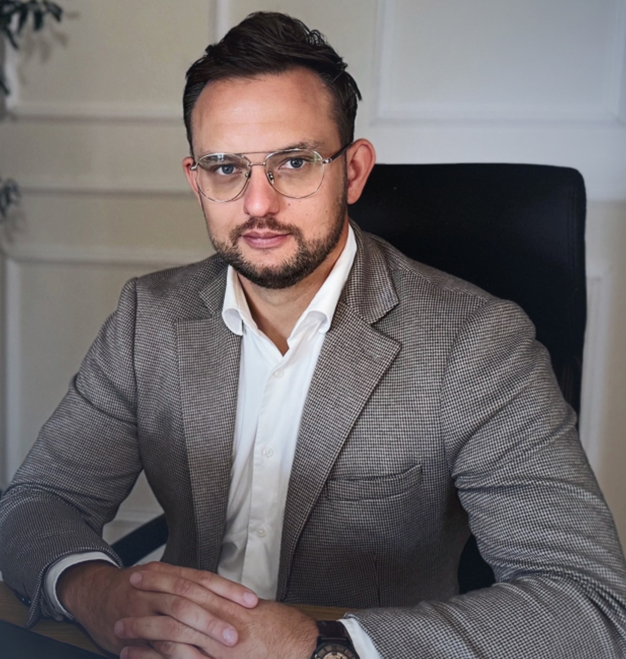 Portret mężczyzny w okularach i marynarce siedzącego za biurkiem. Widoczny zegarek na ręku, stonowana kolorystyka, profesjonalny wygląd. Neutralne tło.